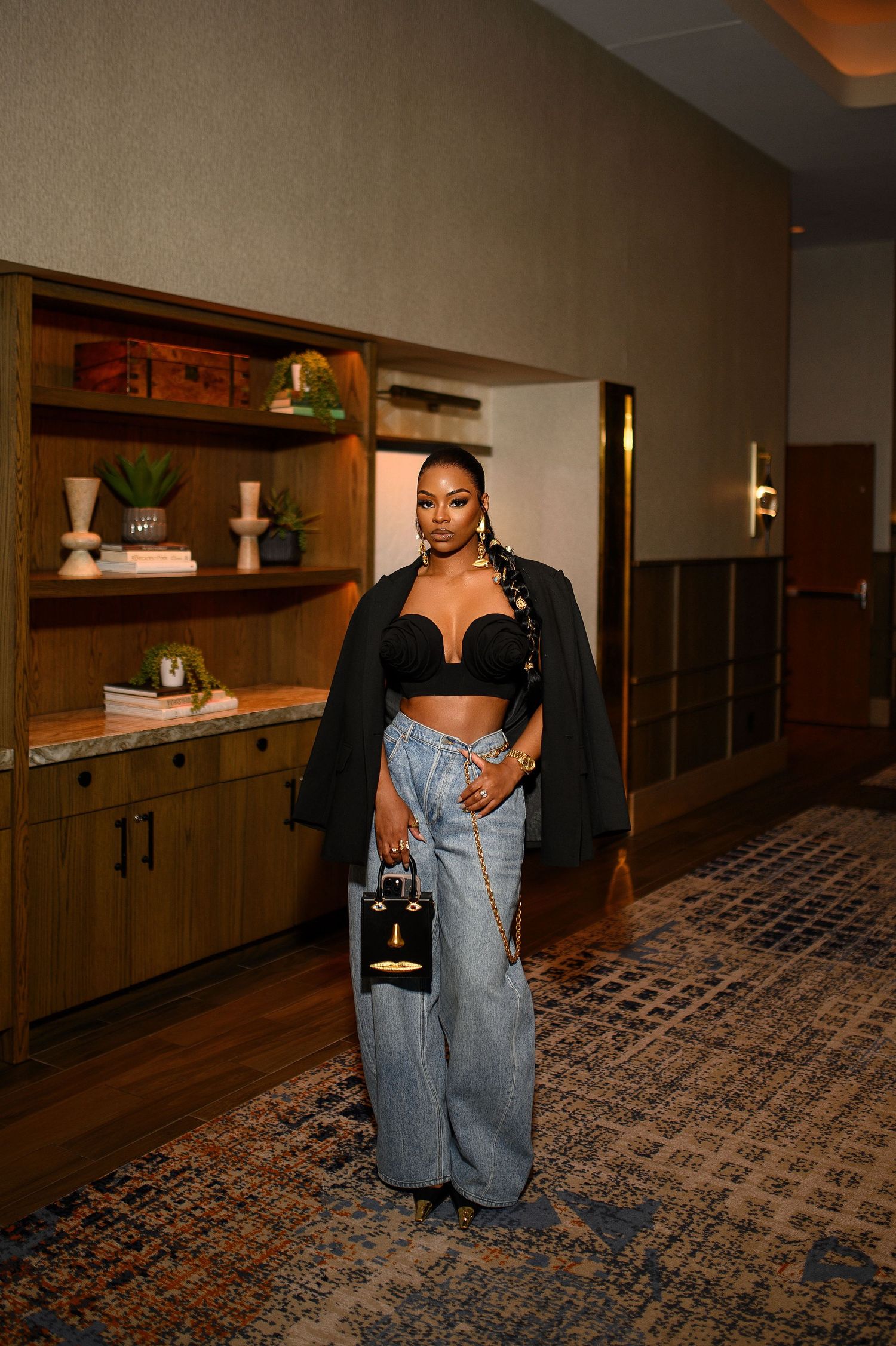 Woman in black crop top, blazer, and wide-leg jeans holds a designer bag in a hotel hallway.