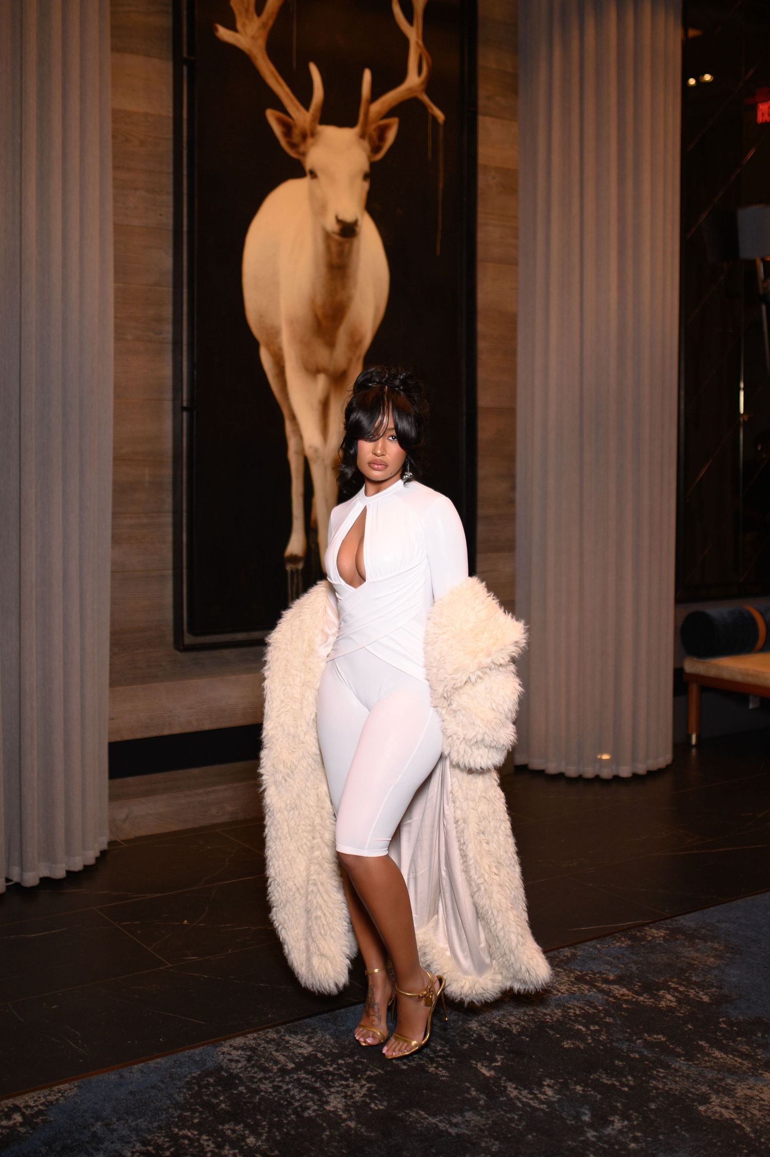 Fashion model in white cutout bodysuit and cream fur coat poses before deer painting.