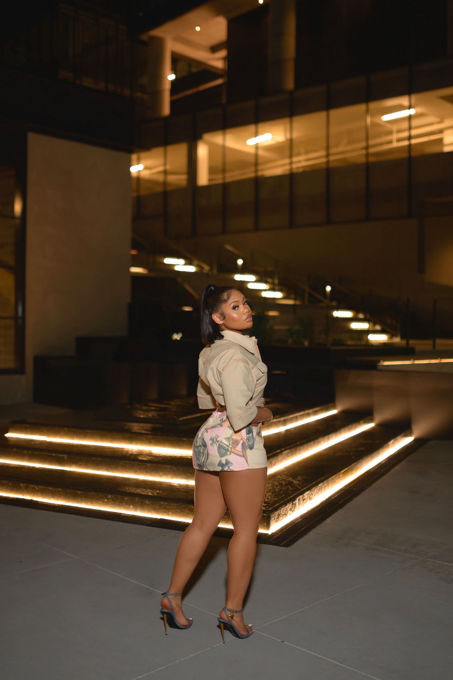 Woman in beige jacket and floral shorts poses in front of an illuminated modern building at night.
