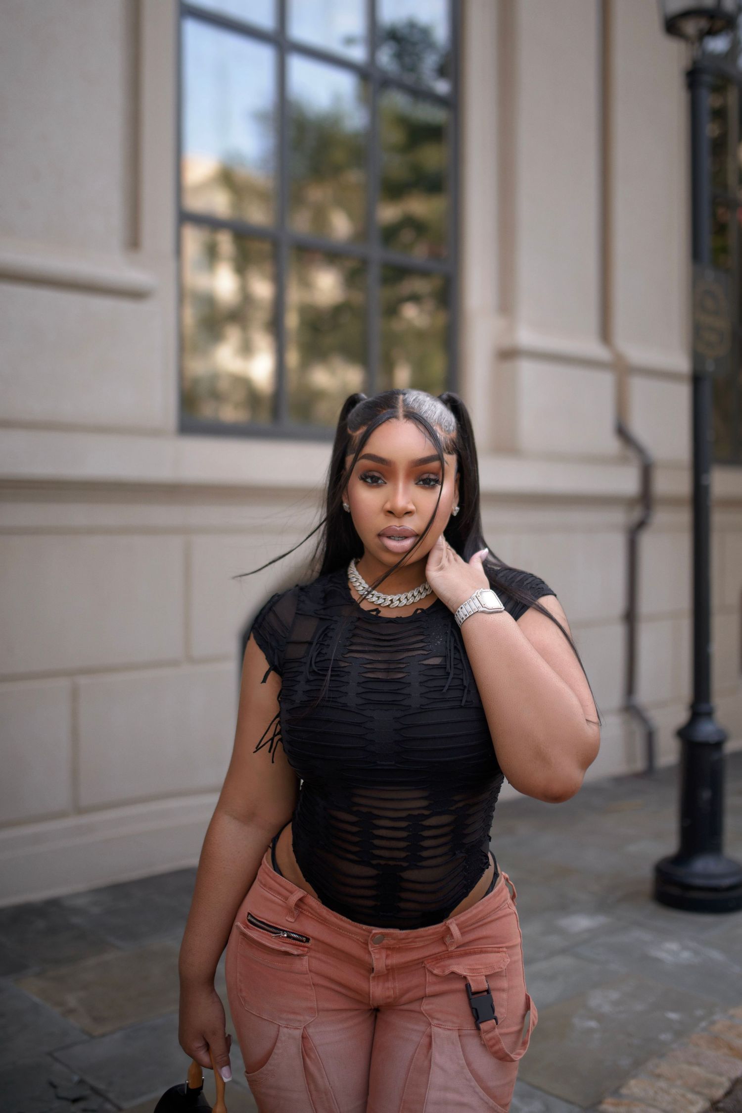 Young woman in black sheer top and pink cargo pants poses outdoors near a beige stone building.