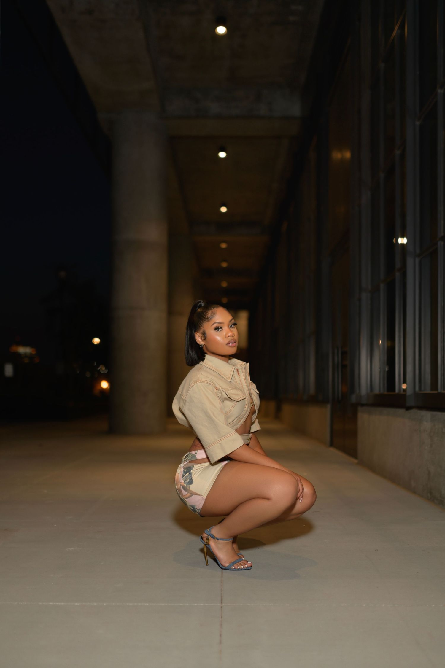 Woman in shorts and heels crouching in nighttime outdoor corridor.