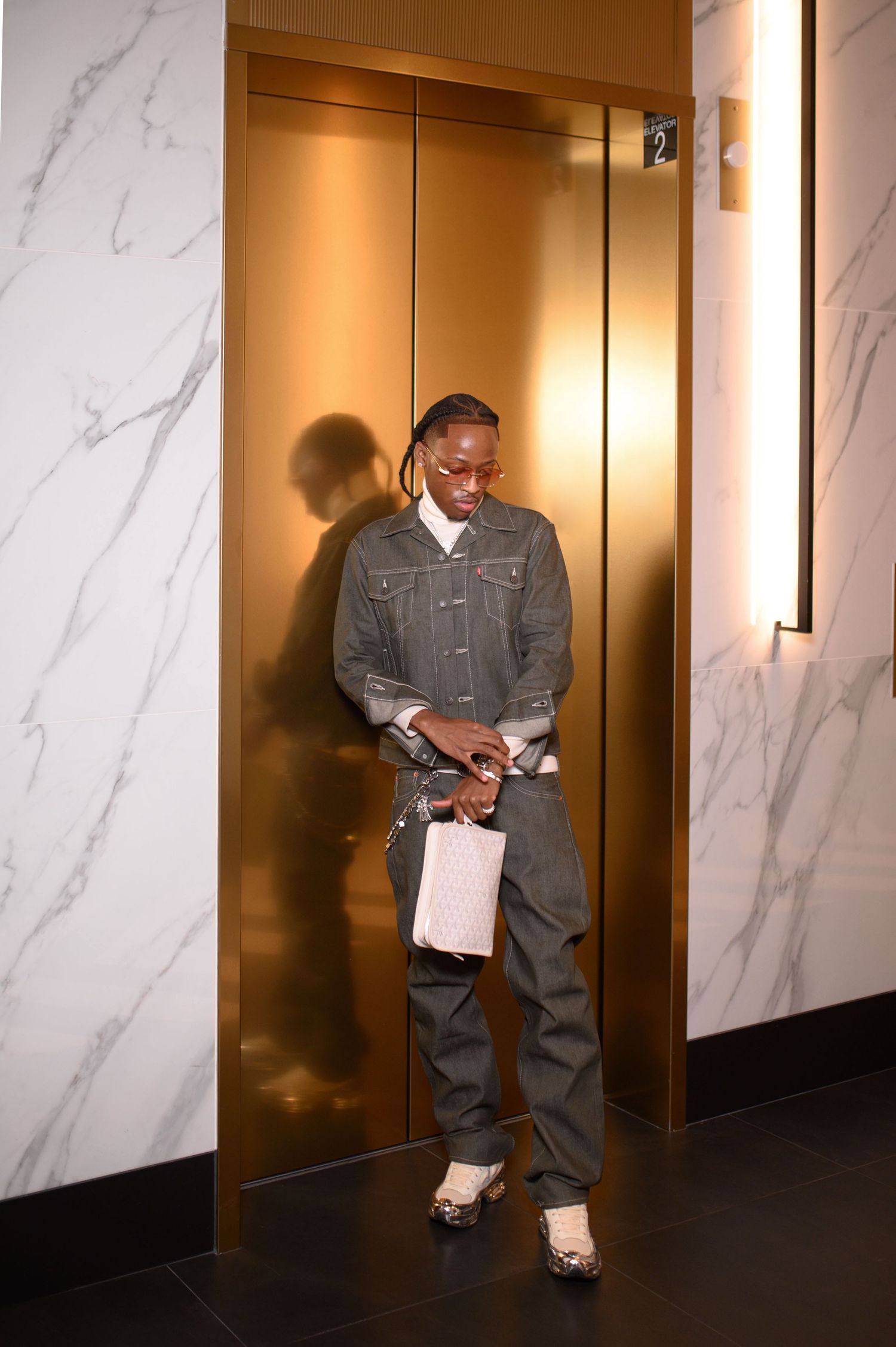 Stylish man in gray denim jacket smiles while leaning against gold elevator doors.