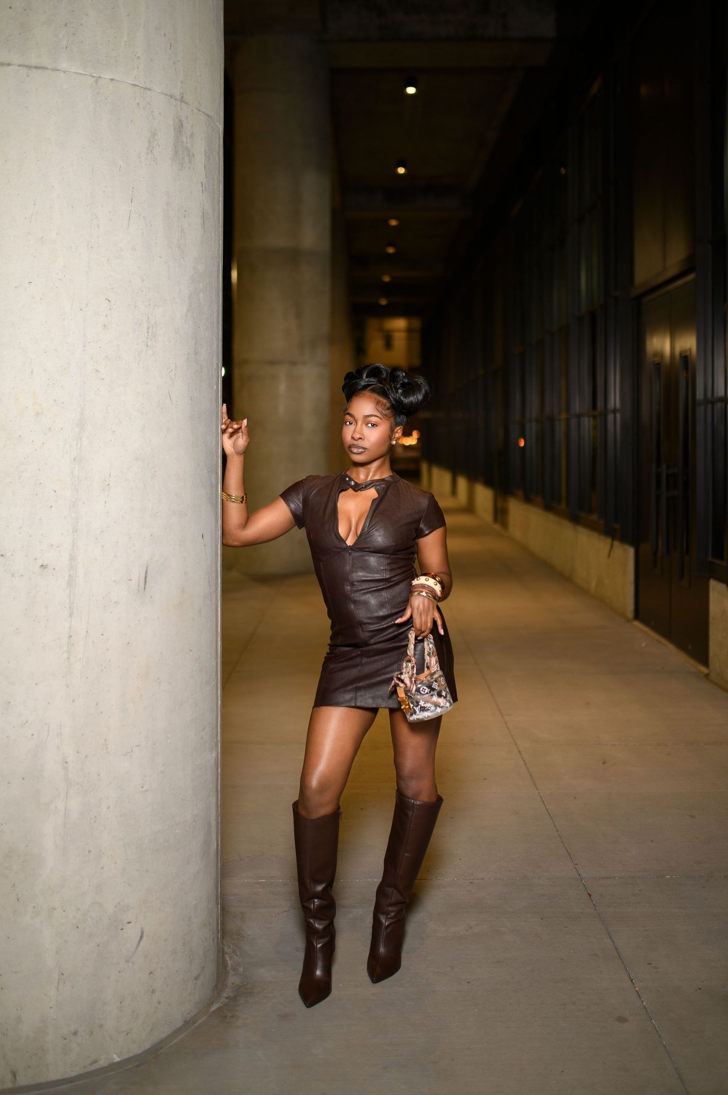 Woman in brown leather mini dress leaning on column indoors.