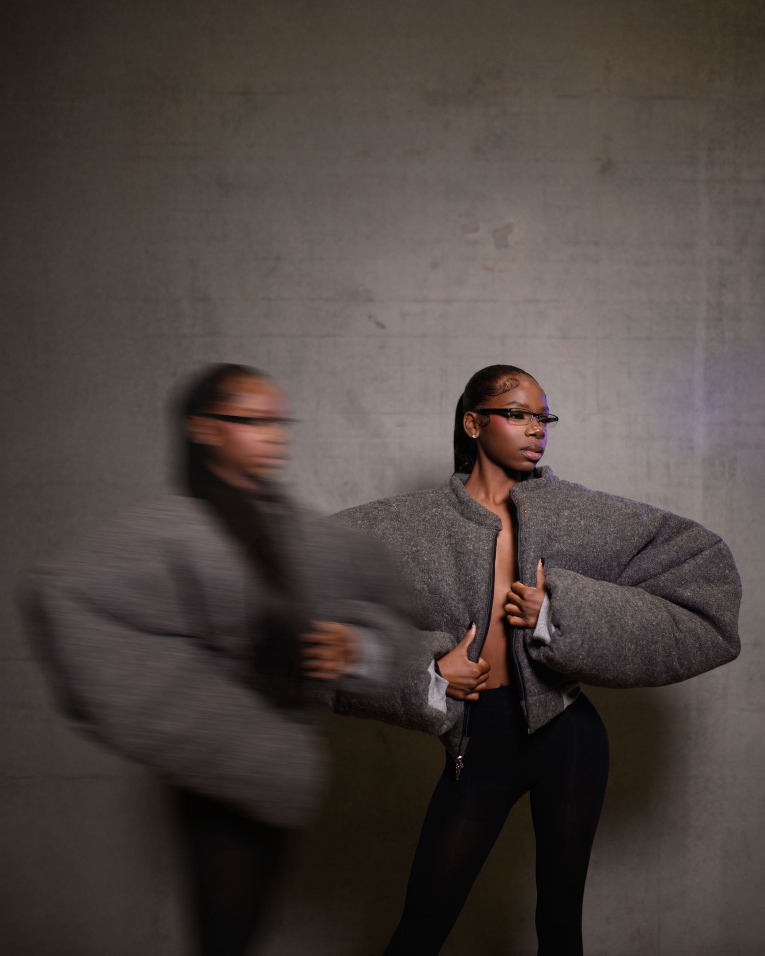 Dynamic double exposure of model in oversized gray puffer jacket against concrete wall.