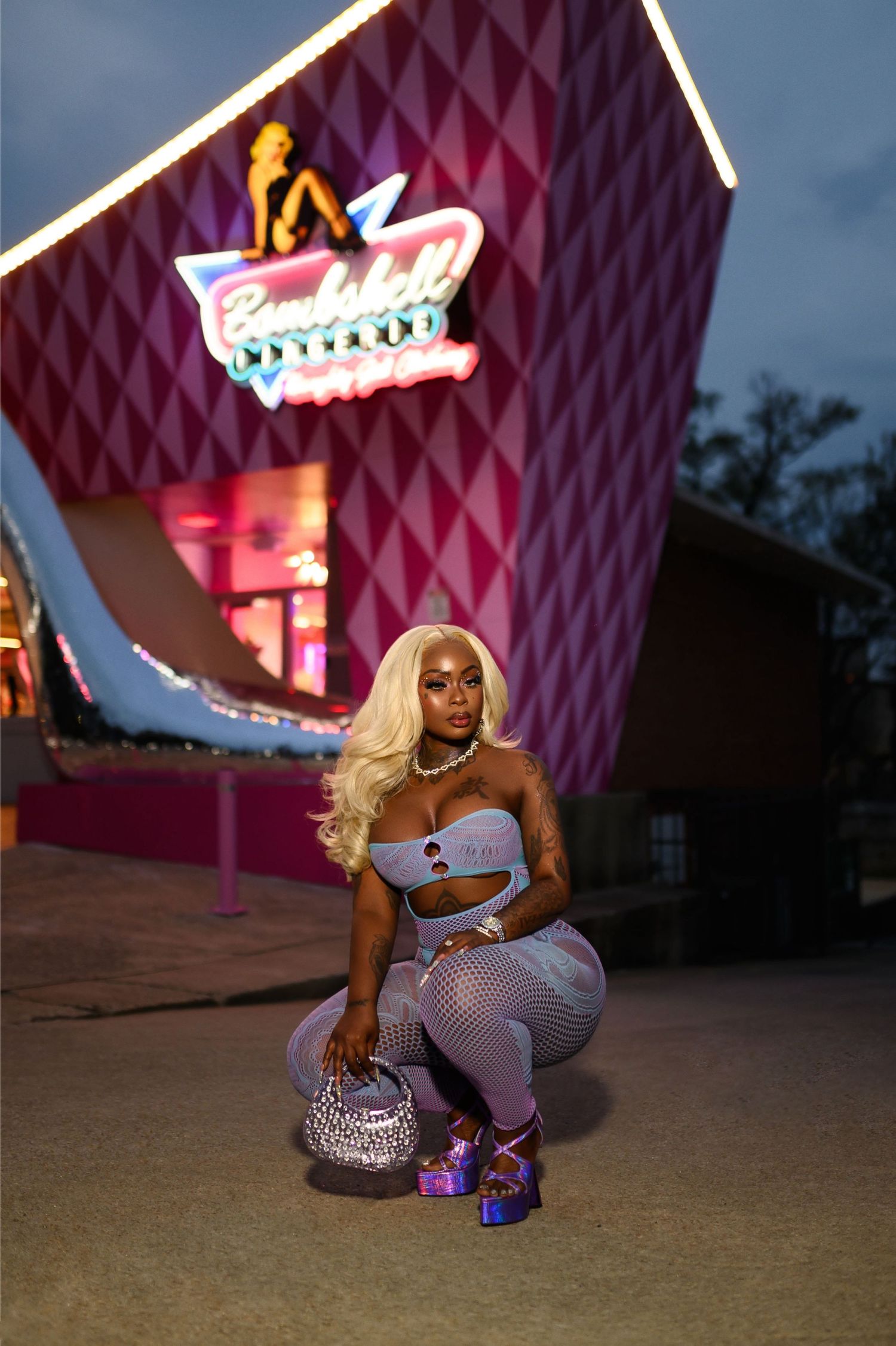 Blonde woman in silver crop top and fishnet pants crouching outside a pink neon-lit building at night.