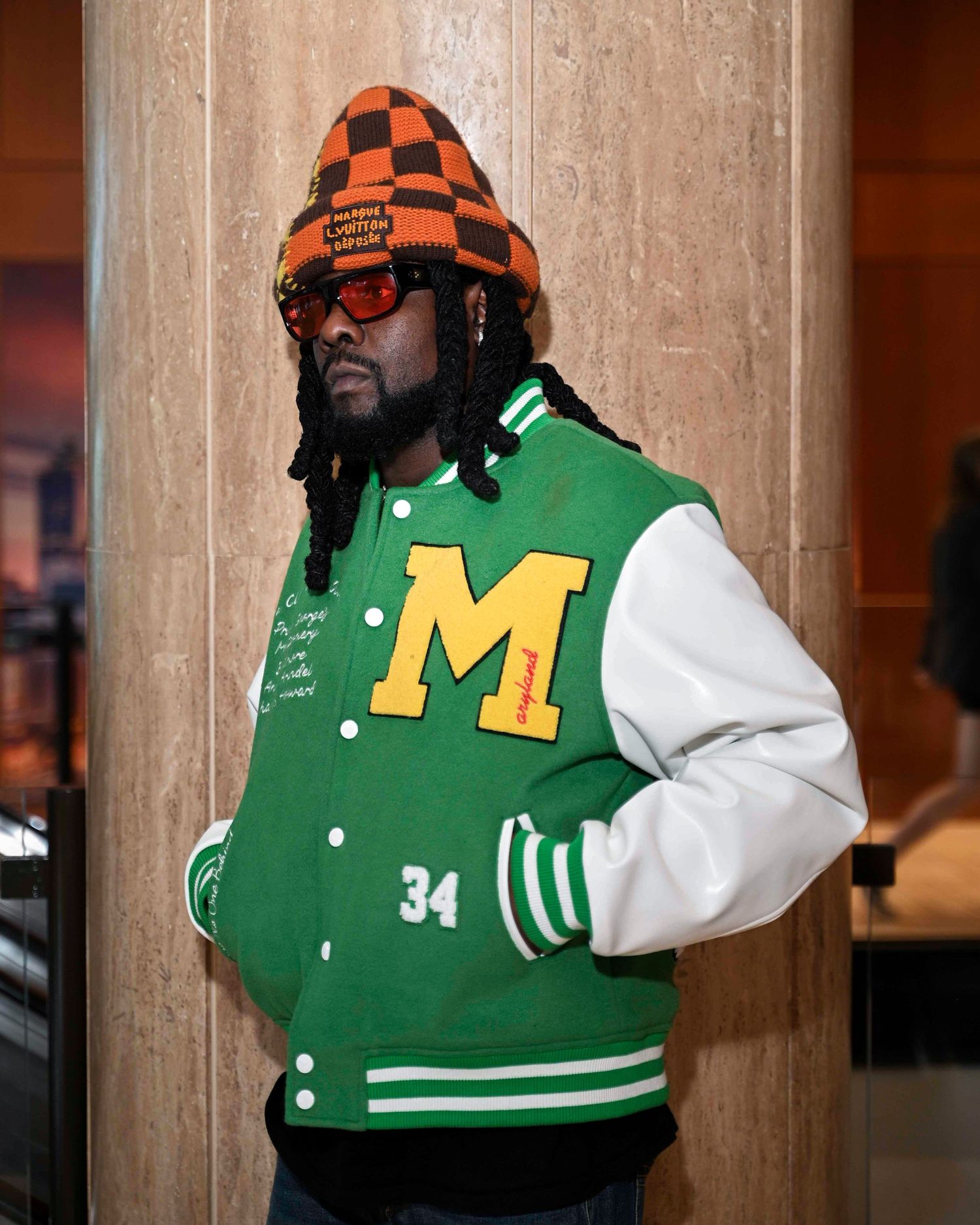 Man wearing green M varsity jacket and orange checkered beanie posing indoors.