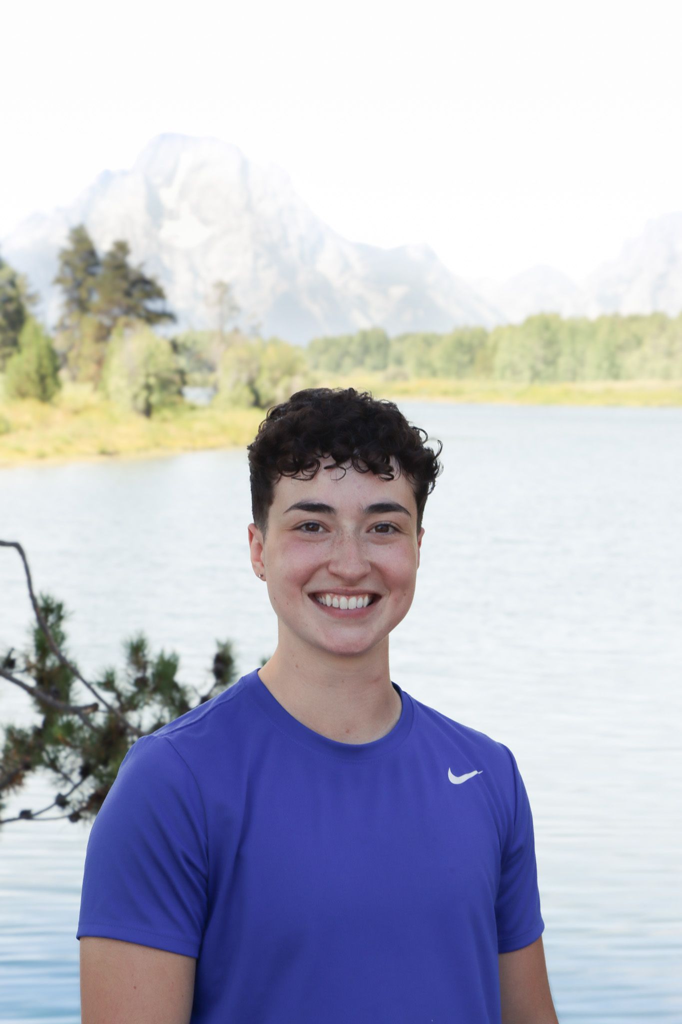 A person in a blue Nike t-shirt smiles while standing in front of a scenic lake and mountain landscape.