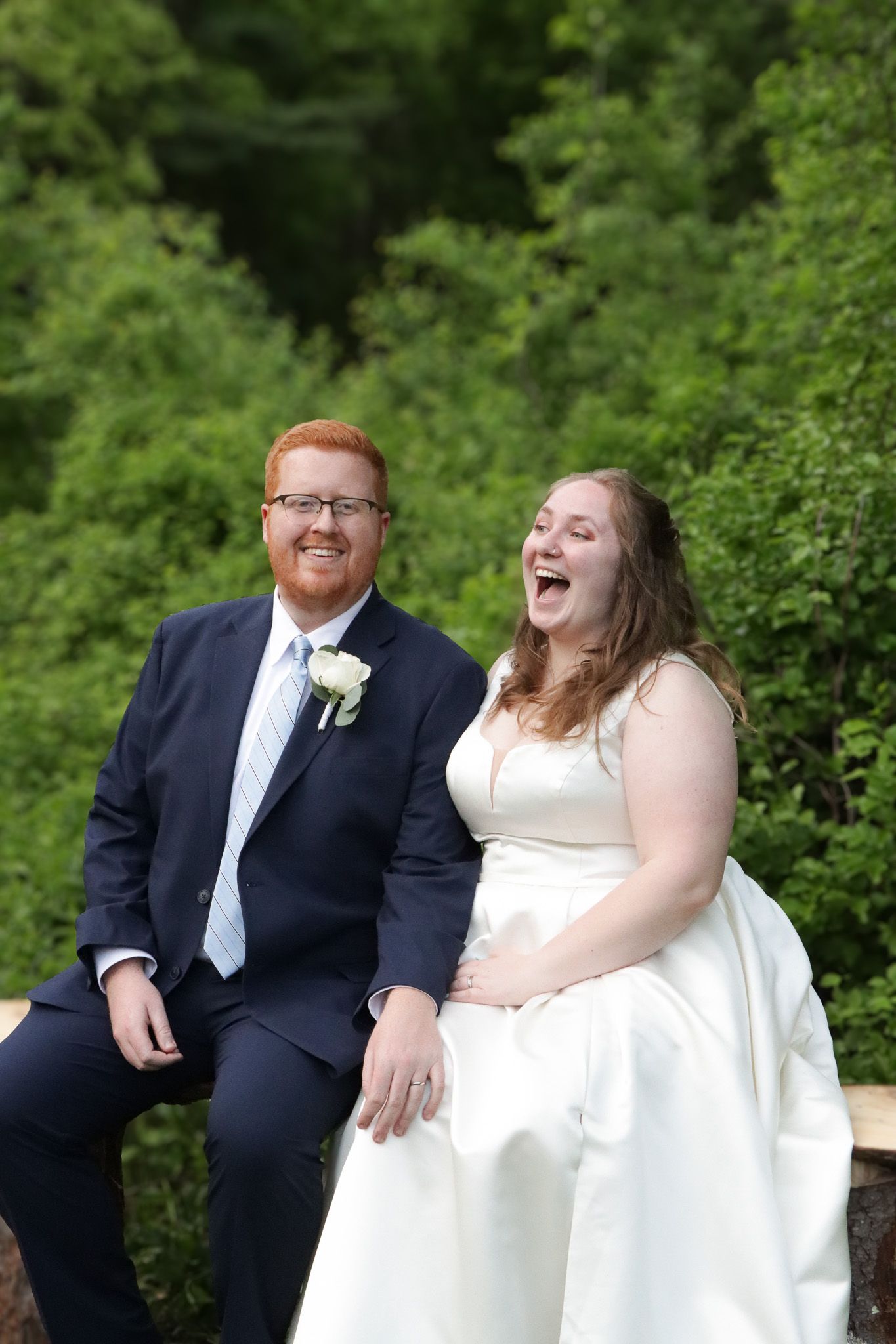 A happy couple shares candid laughing moments during their wedding in a lush green outdoor setting.