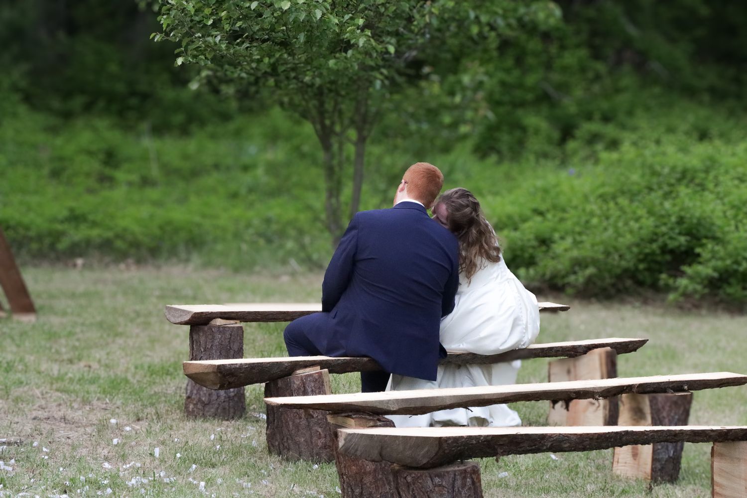 Bride and groom have an intimate moment together sitting on a wooden bench in their wedding ceremony space.