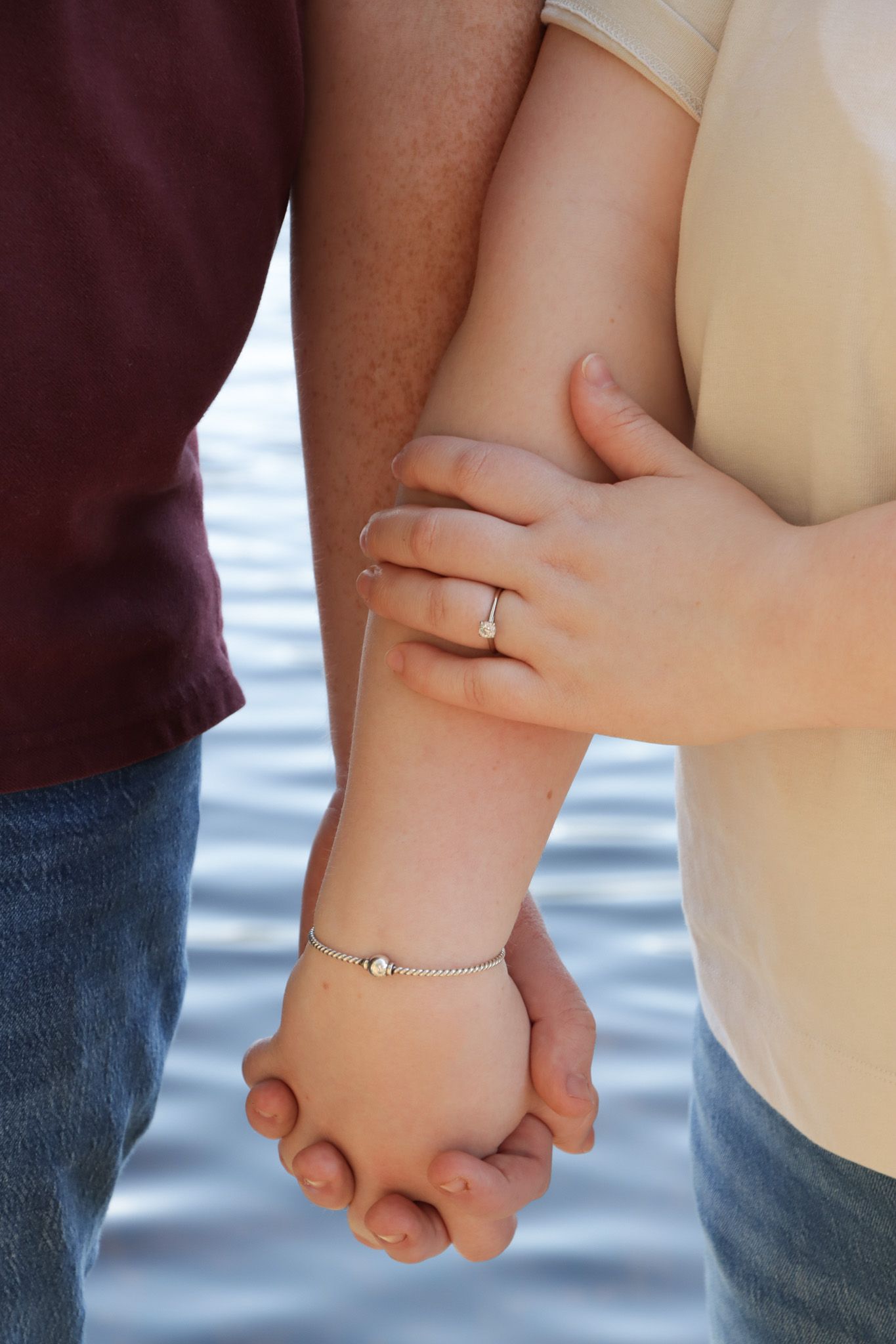 Close-up of two people holding hands with fingers interlocked against a blurred lake water background and close up of engagement ring.