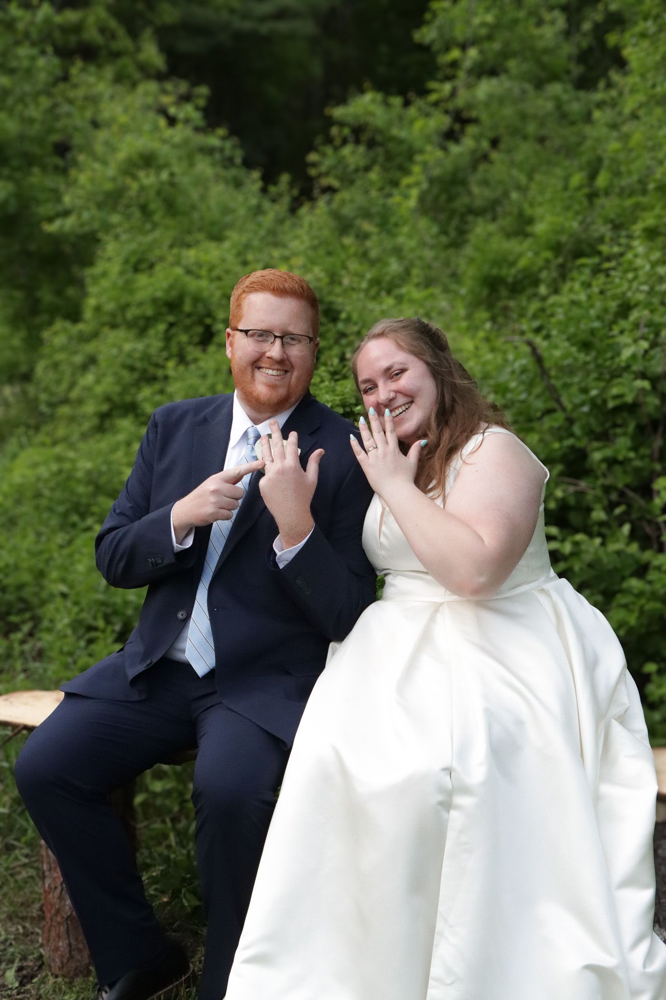A newly married couple show off their wedding rings surrounded by a lush green background.