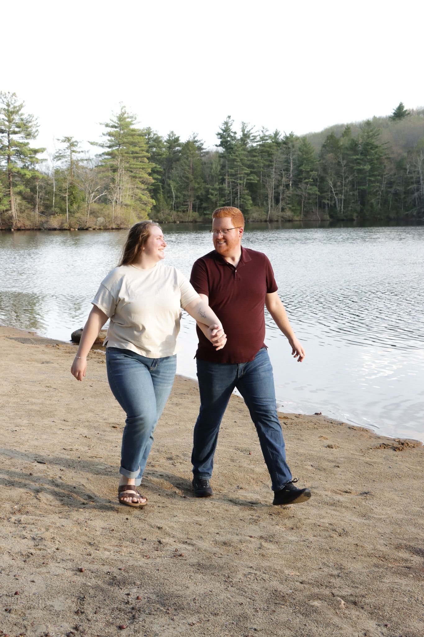 A couple walks together along a sandy beach beside a serene lake surrounded by pine trees on a calm day at a Massachusetts park.