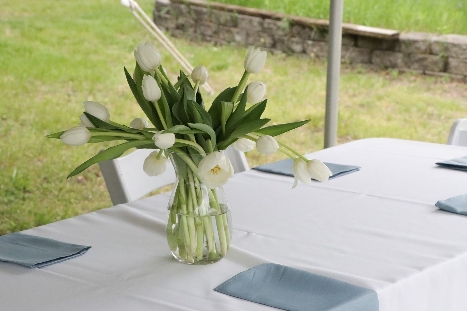 White tulips in a glass vase sit on an outdoor white clothed table for wedding guests.