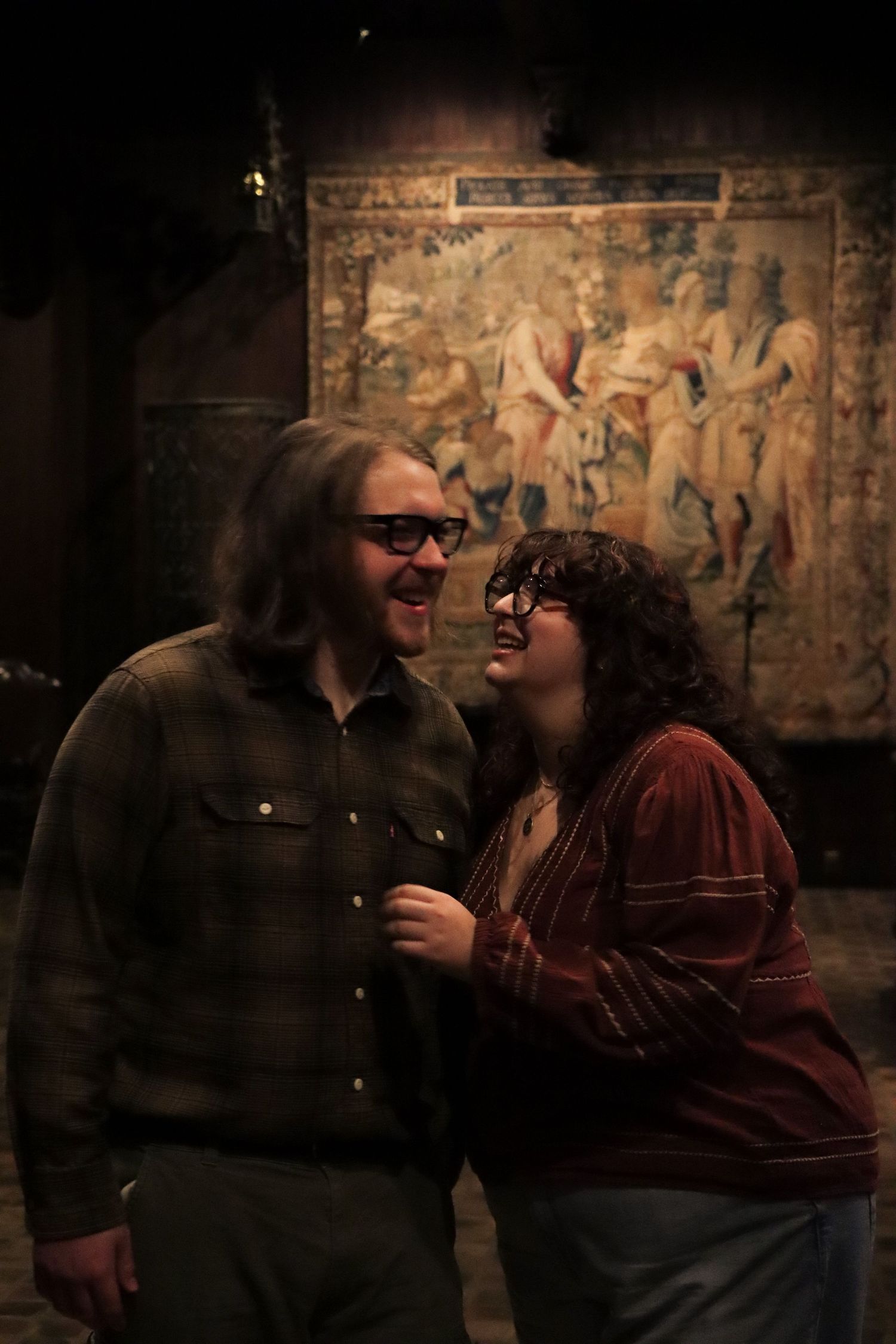 A couple shares a joyful moment in a dimly lit room with vintage tapestries on the wall behind them.