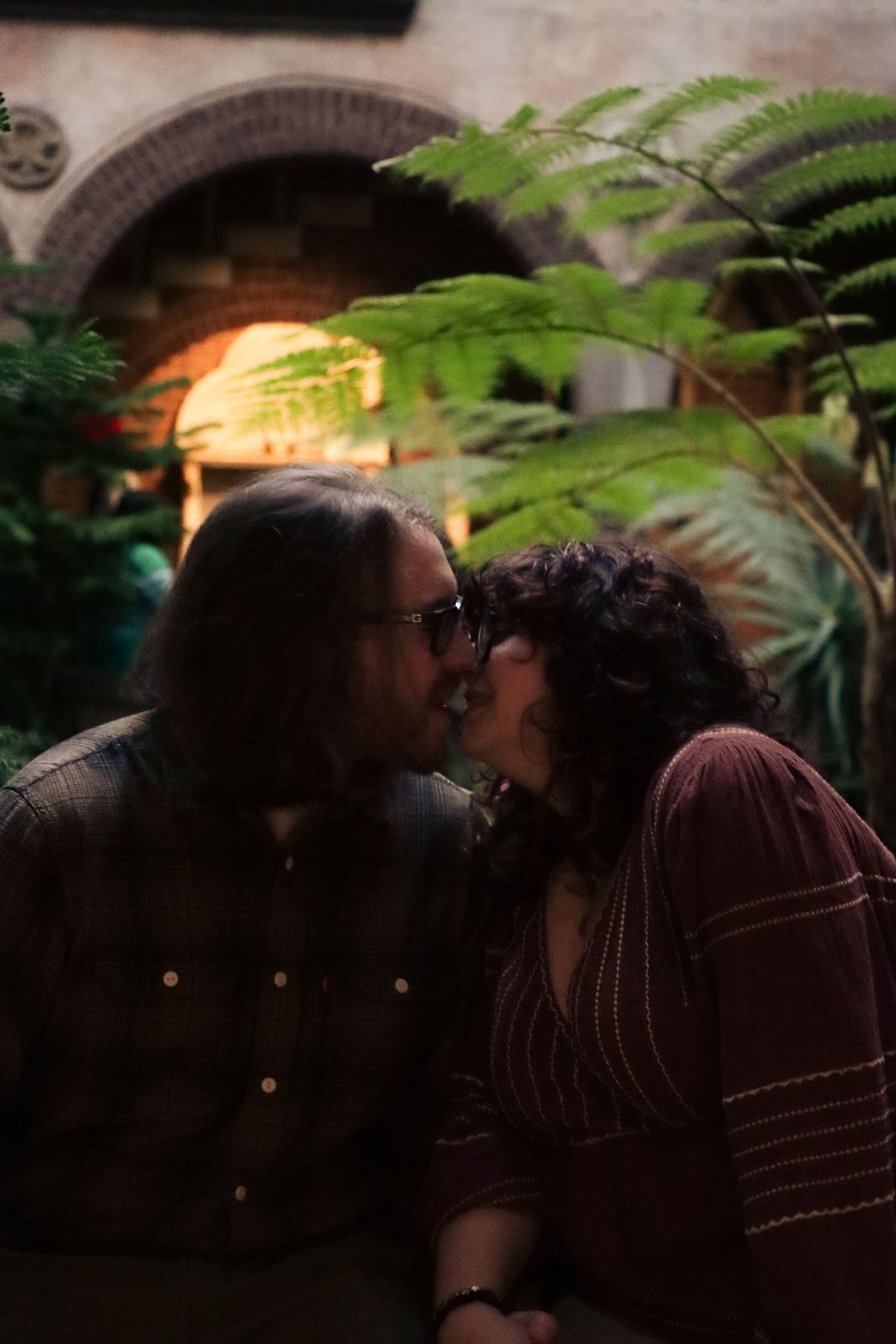 A couple shares a romantic kiss under fern leaves and warm lighting at a Boston Massachusetts museum.