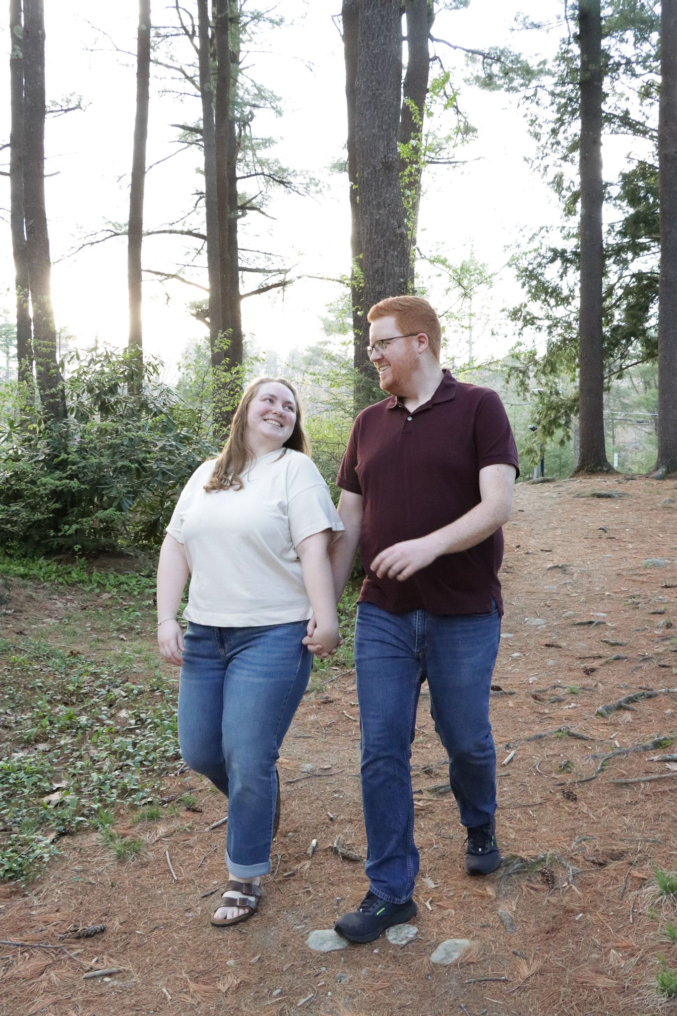 A happy couple walks down hill amongst tall trees and a bright sky in a Massachusetts park.