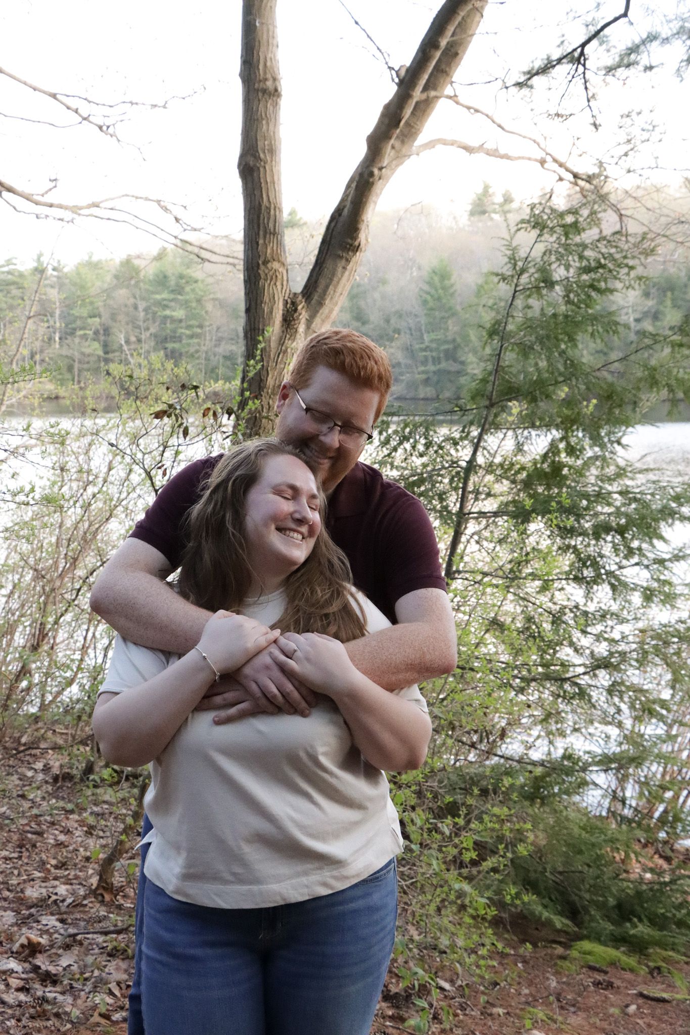 A couple shares a joyful embrace while standing near a lake surrounded by trees at a Massachusetts park.