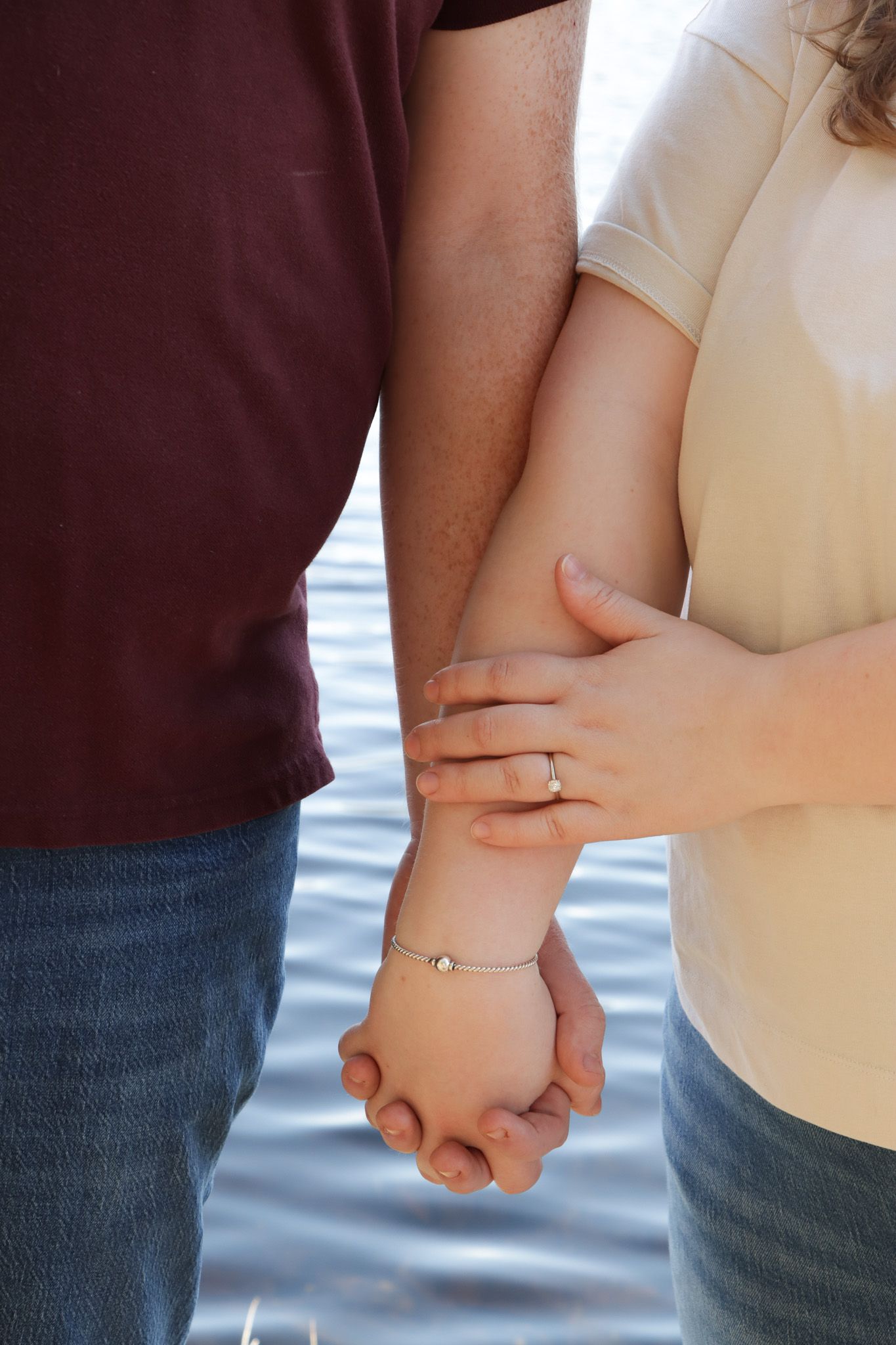 Close-up of two people holding hands with fingers interlocked against a blurred lake water background and close up of engagement ring.