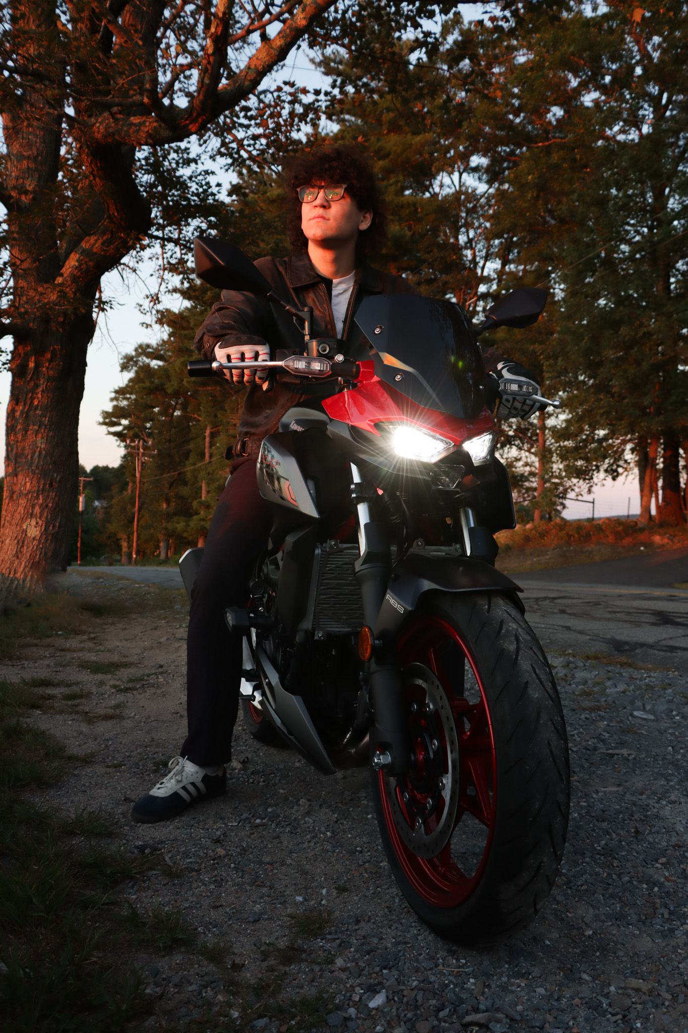 A motorcyclist on a black and red sport bike poses at sunset in a wooded area with the bike's headlight illuminated.