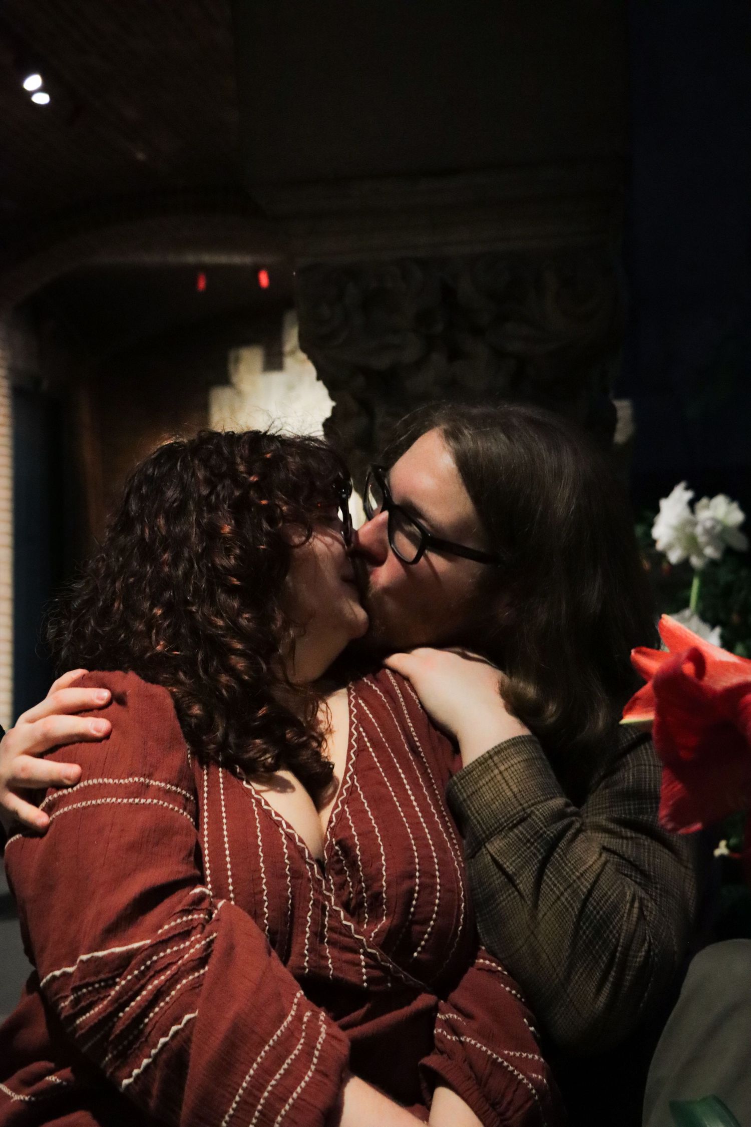 Two people sharing an intimate kiss in a dimly lit room with red flowers visible in the background.