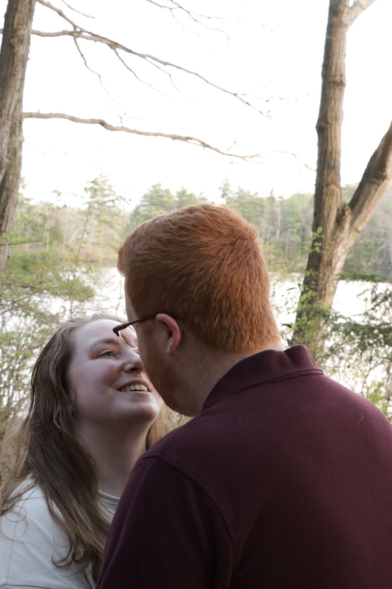 A couple shares an intimate moment outdoors against a backdrop of bare trees and natural scenery at a park in Massachusetts.