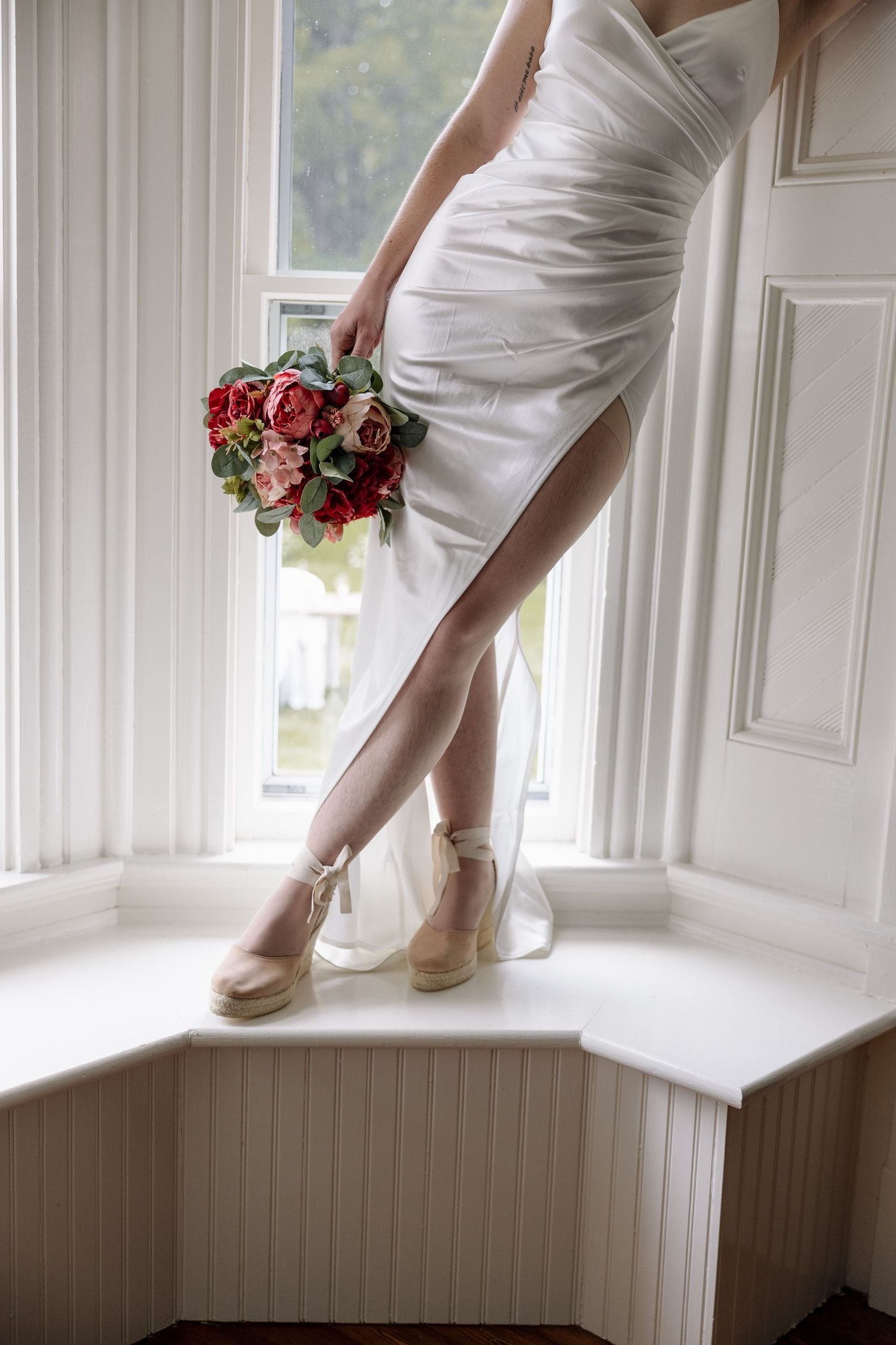 Bride in white dress holding red rose bouquet, standing on window seat wearing nude wedge heels.