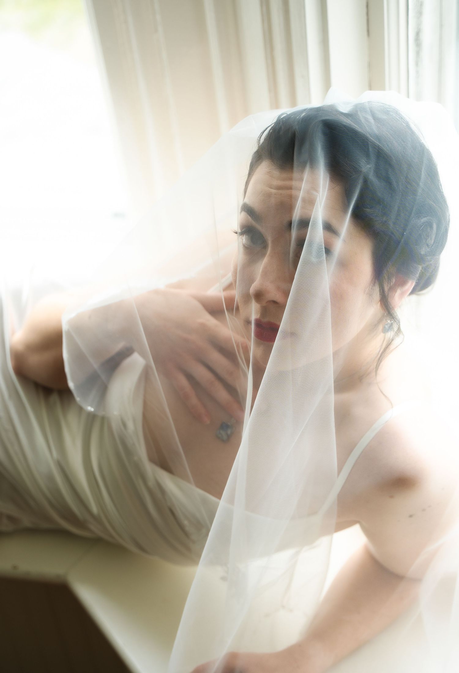 Bride in white wedding dress and veil gazing thoughtfully by a bright window in a serene bridal portrait.