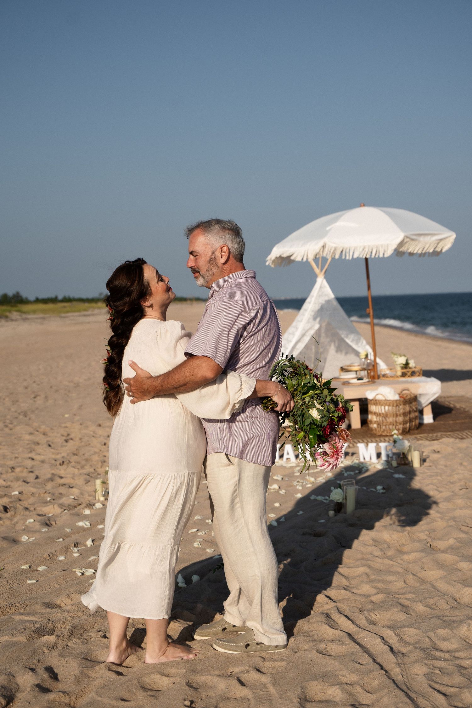 Couple embracing on a sandy beach near a white umbrella and picnic setup on a clear sunny day.