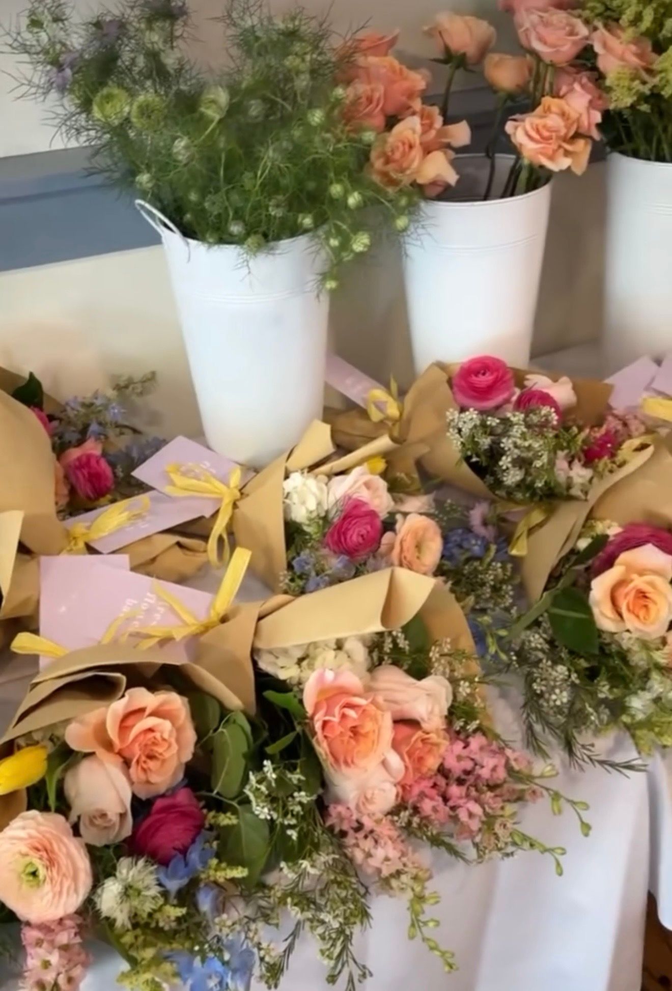 Colorful spring flower bouquets wrapped in kraft paper with yellow ribbons displayed on a table with white vases of pink roses.