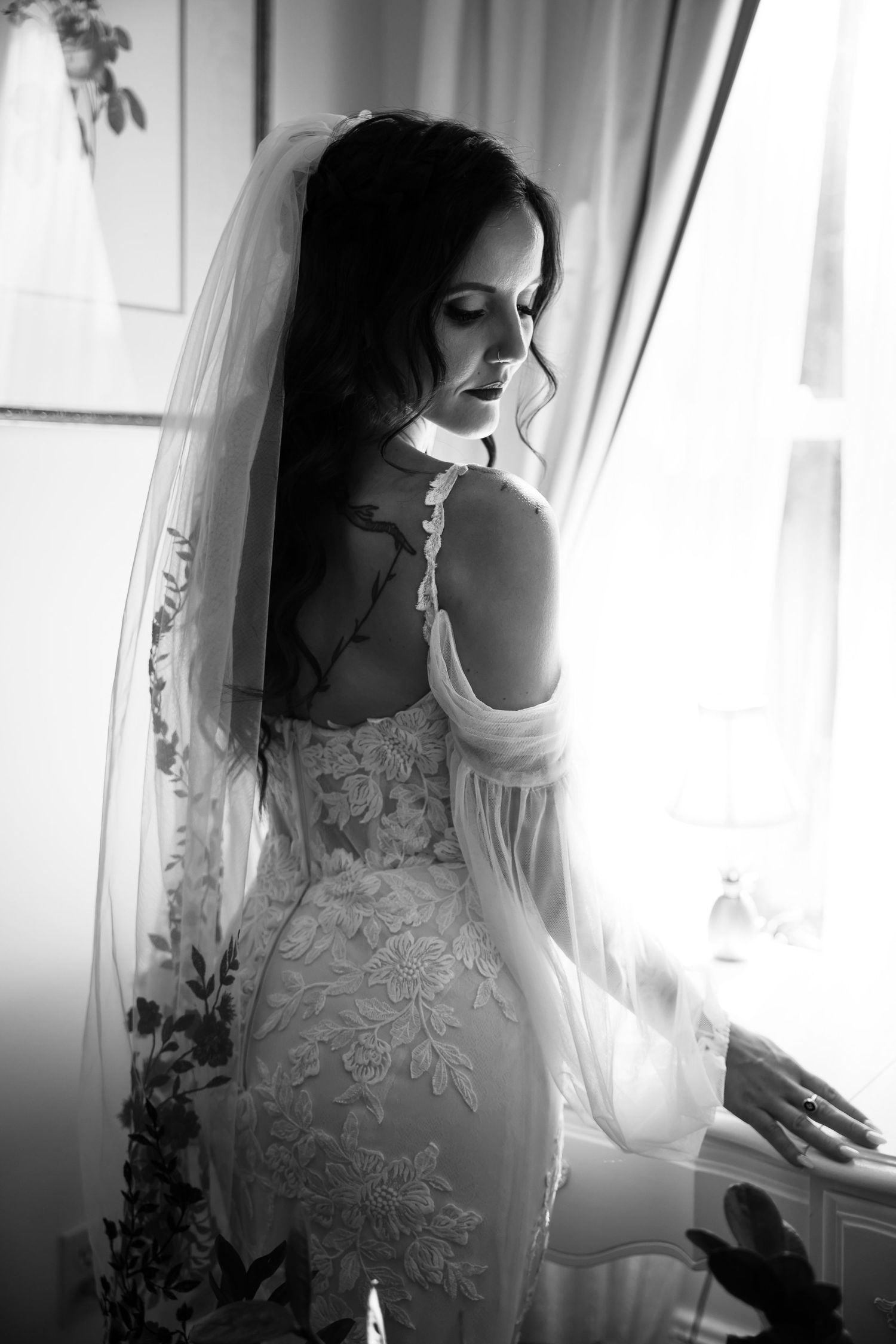 Black and white photo of a bride in lace wedding dress and veil standing by a window looking down.