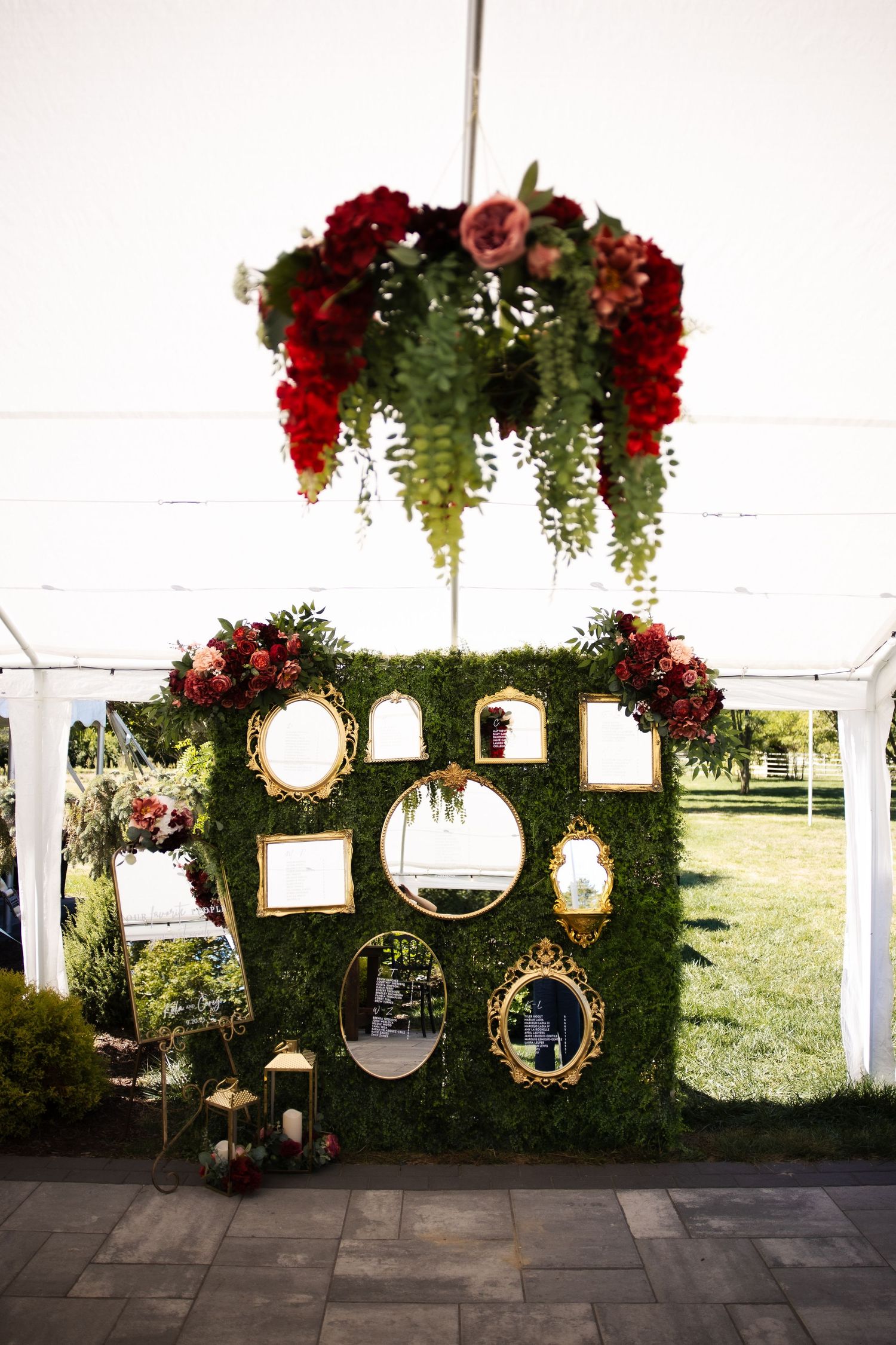 Elegant wedding backdrop with gold-framed mirrors on a lush green moss wall, adorned with red floral arrangements under a tent.