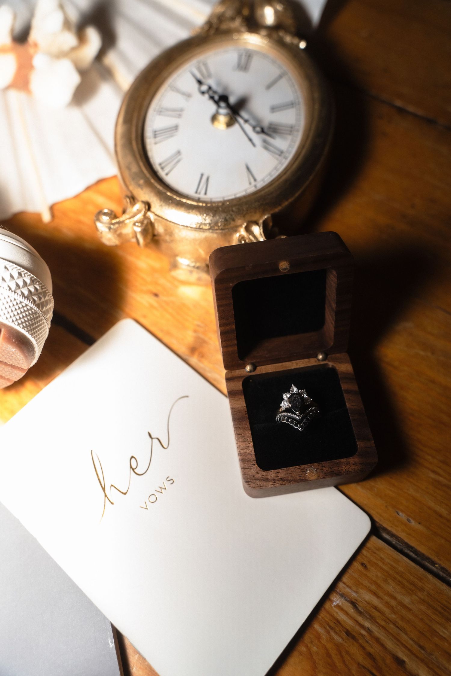 Elegant wedding flat lay with diamond ring in wooden box, her vows book, and vintage gold clock on wood table.