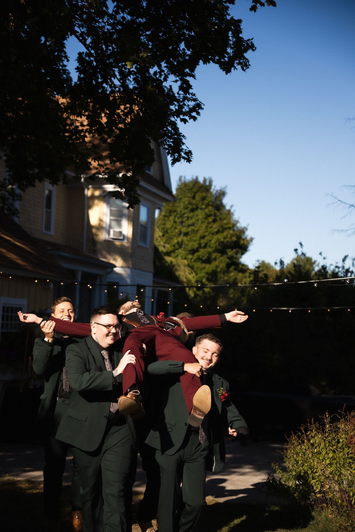 Groomsmen in dark suits lift groom in burgundy tuxedo outdoors at wedding celebration under blue sky.