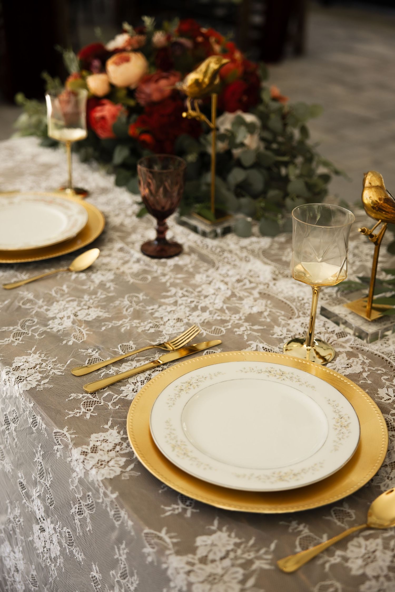 Elegant gold and white place setting on lace tablecloth with floral centerpiece and gold cutlery.