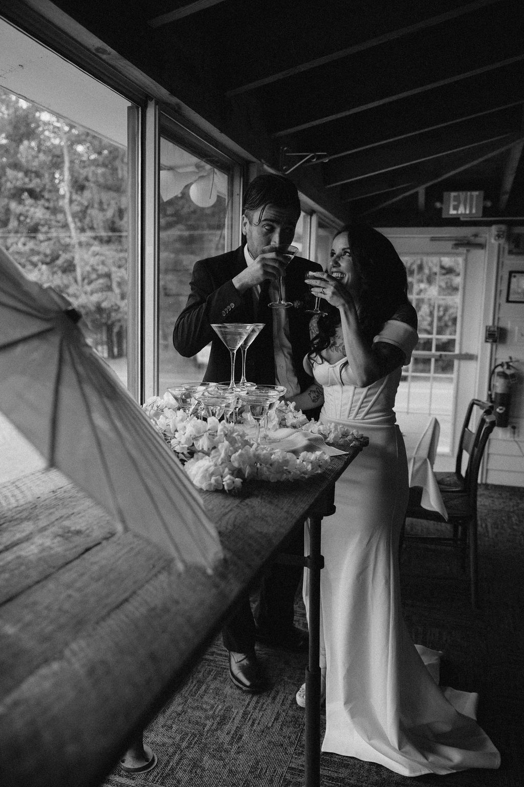 Black and white photo of a bride and groom toasting with cocktails at a table decorated with flowers inside a rustic venue.