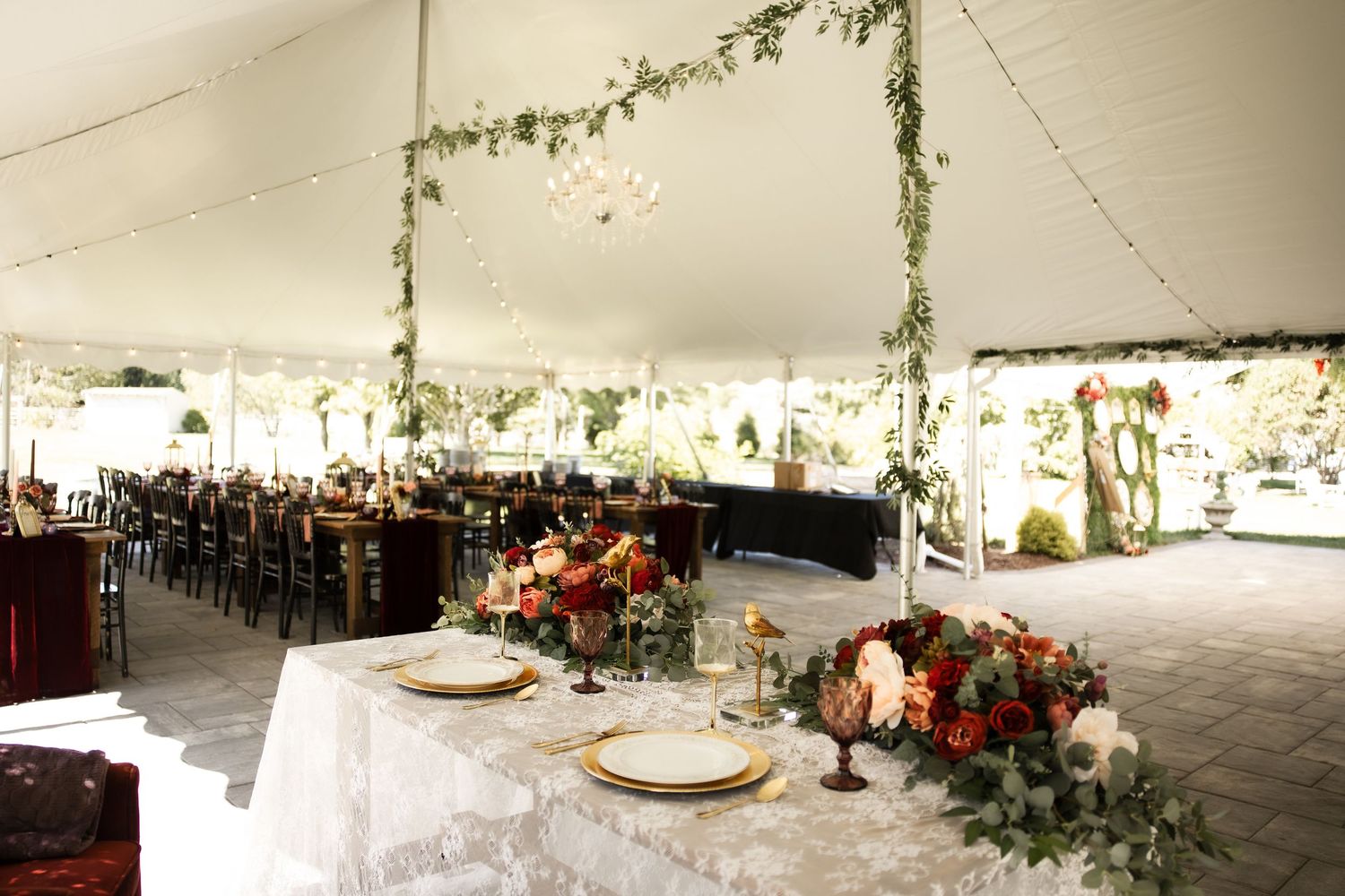 Elegant wedding reception tent with floral arch, chandelier, lace tablecloths, and red floral centerpieces on decorated dining tables.