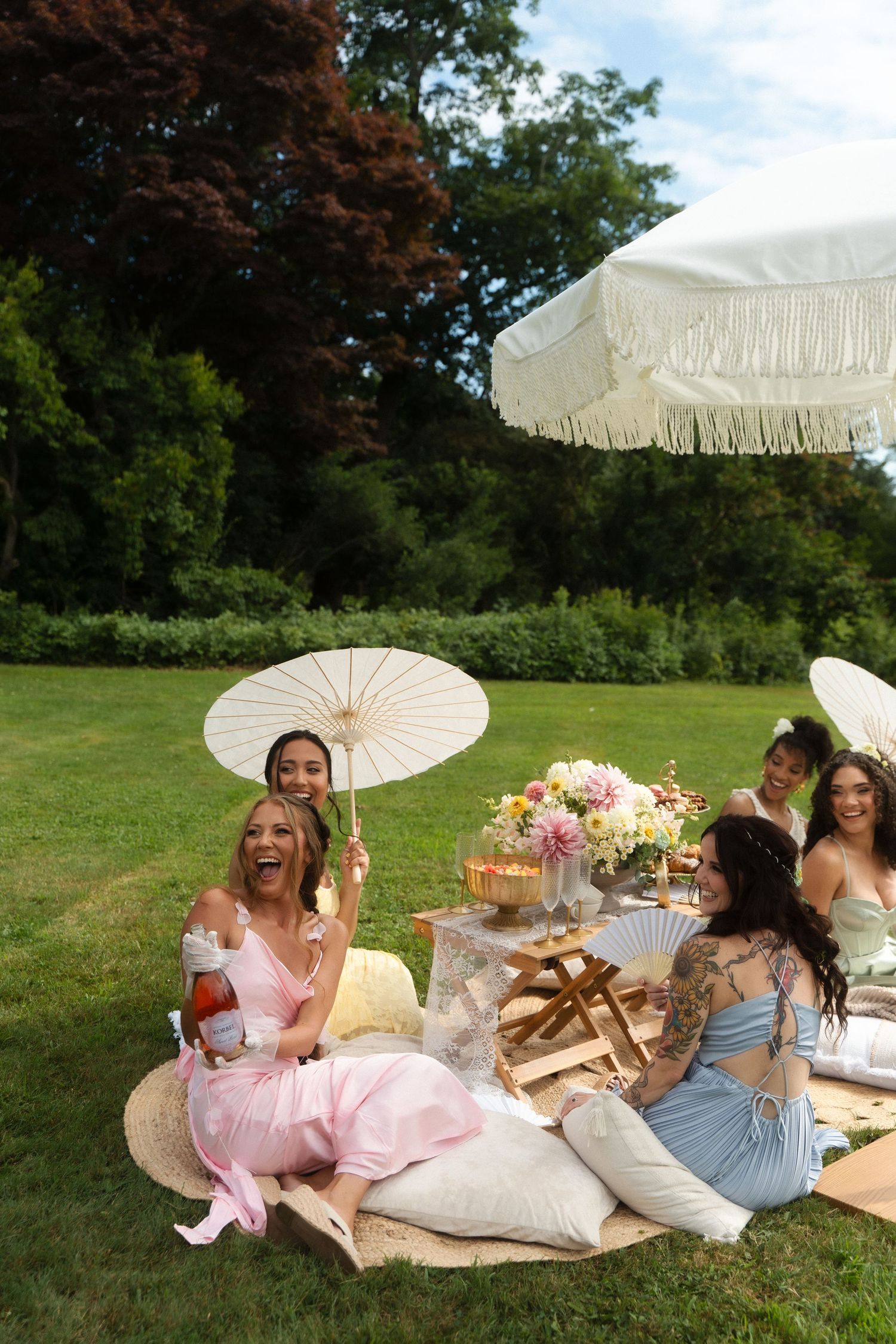 Women in pastel dresses enjoying an outdoor garden picnic with parasols and floral arrangements on a sunny day.
