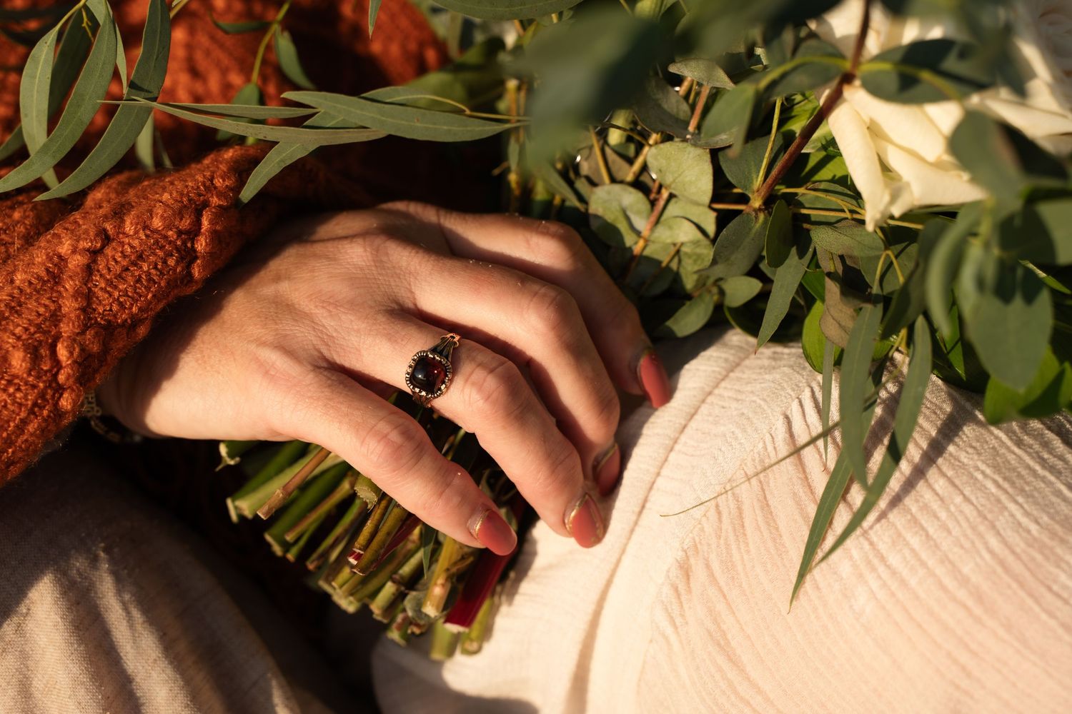 Close-up of a hand wearing a dark gemstone ring, resting on a white dress beside a green eucalyptus bouquet.