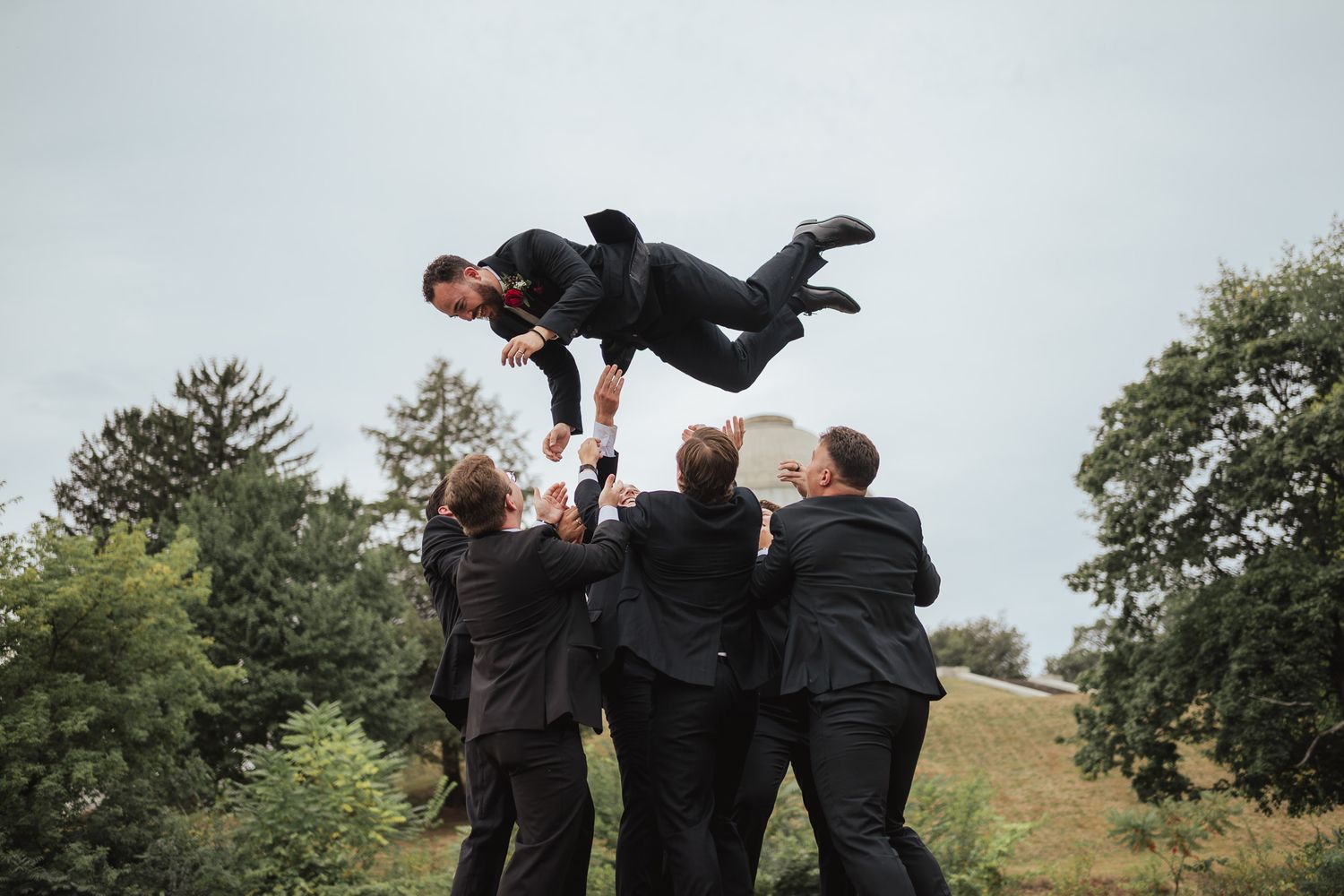 Groomsmen playfully lift and carry each other on a grassy hillside during wedding photos.