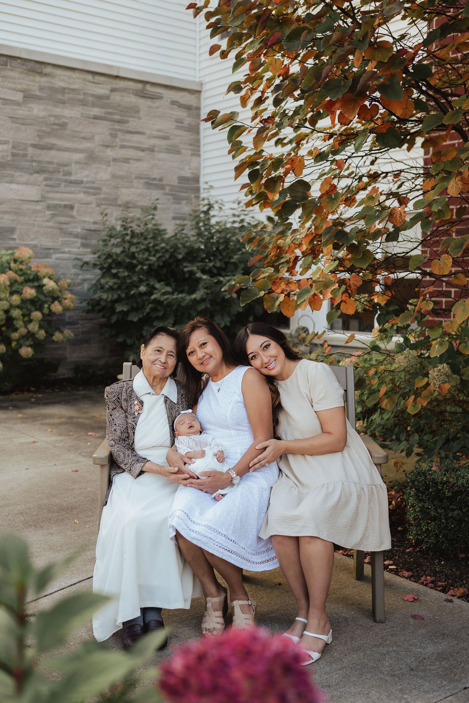 Four generations of women sit on a bench outdoors, smiling together, with a newborn baby in white clothing.