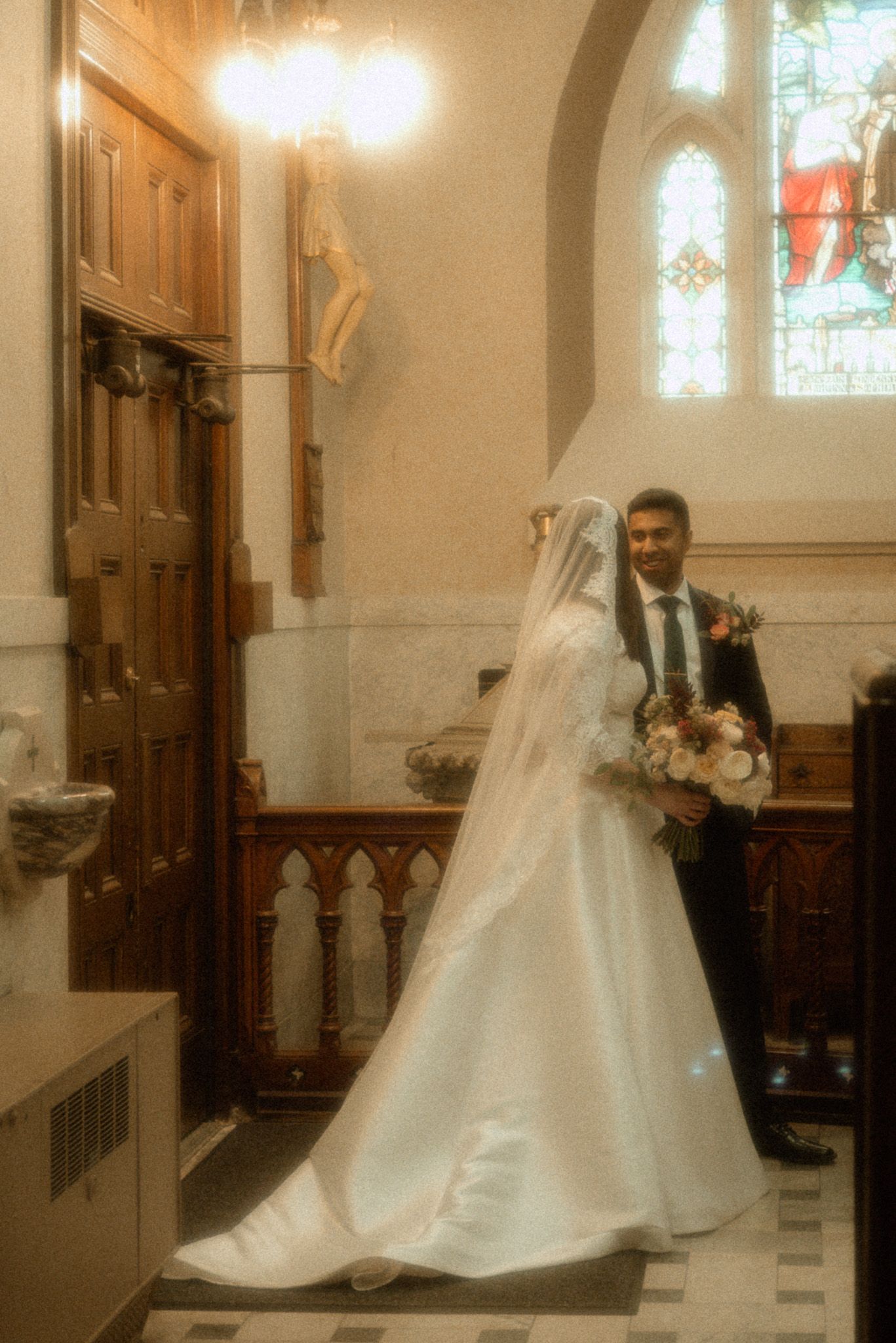 A couple stands in a church on their wedding day, with stained glass windows and wooden details visible in the background.