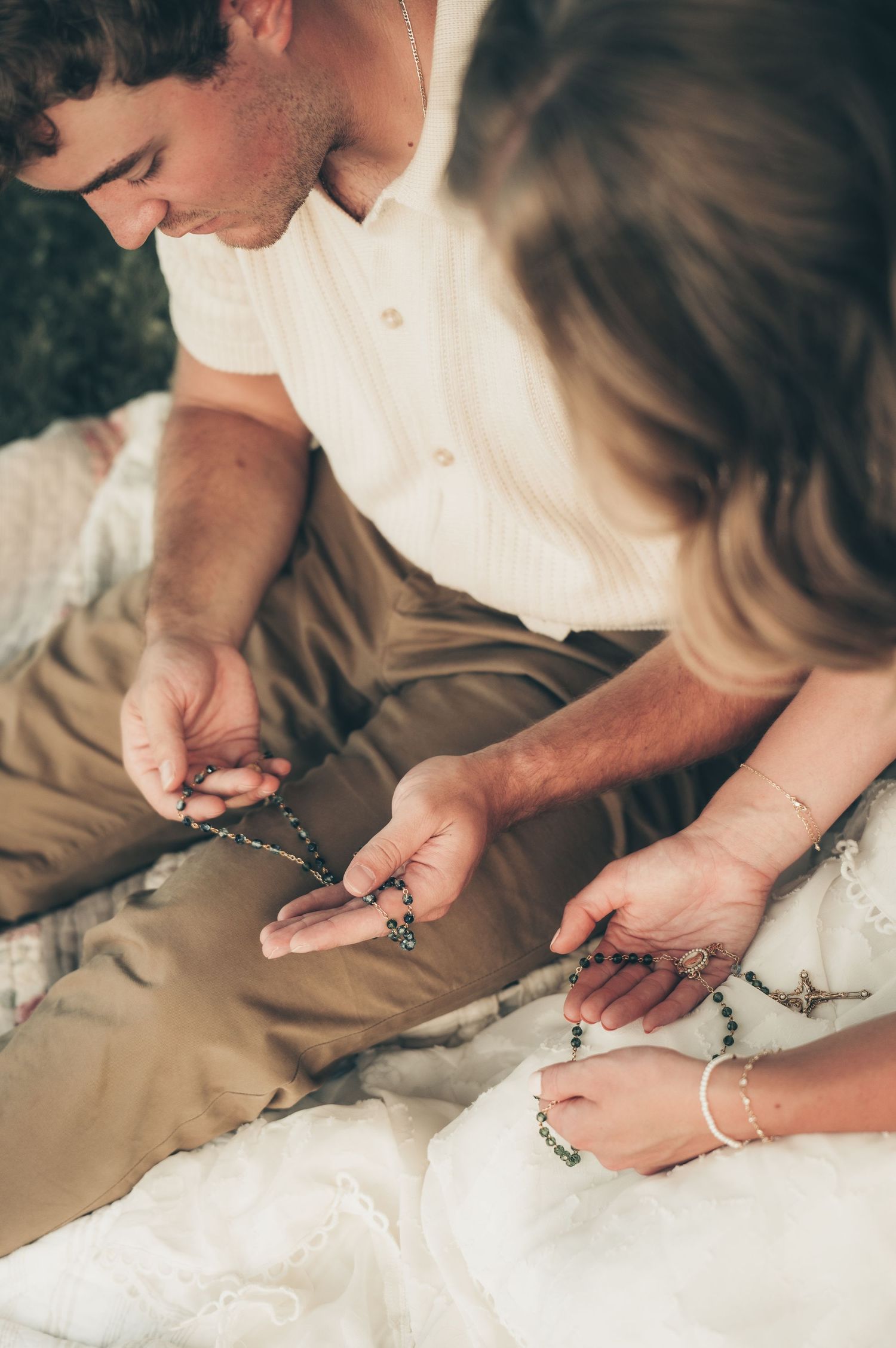 Couple sitting together examining delicate beaded rosary necklaces with cross pendants, wearing casual boho style clothing.