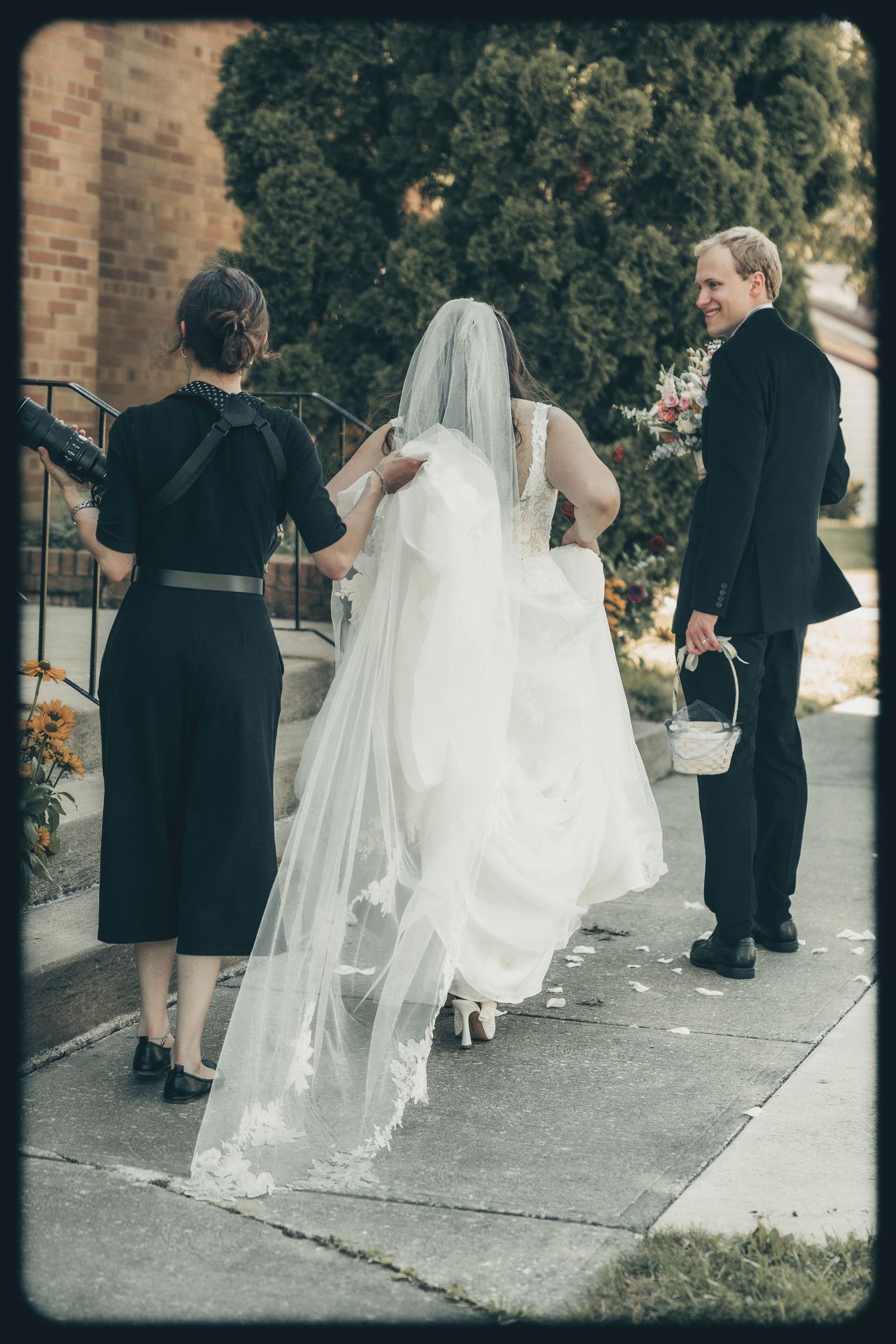 A bride in a flowing white gown and veil walks up church steps with attendants in black dresses.