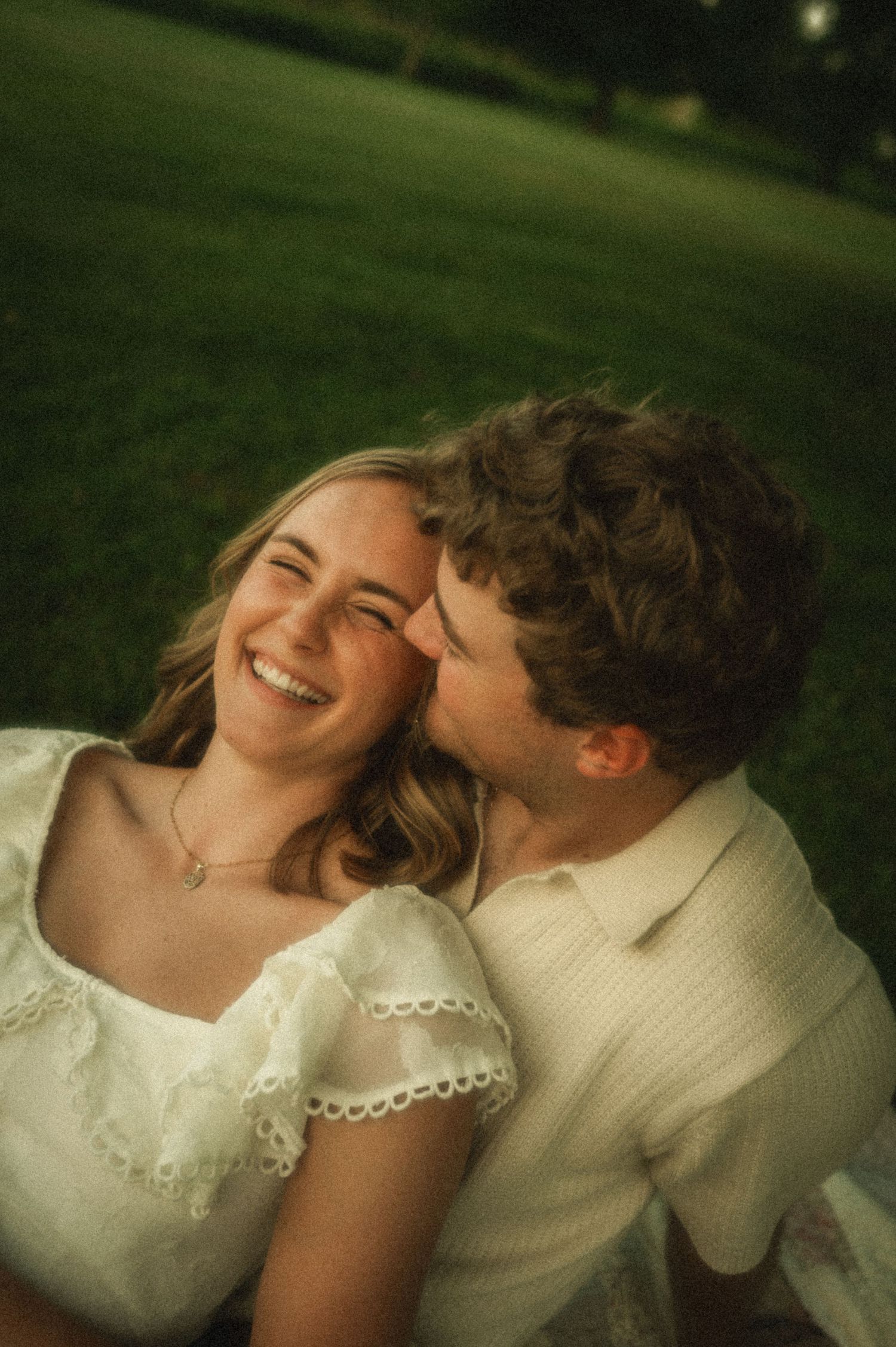 A couple shares a tender moment outdoors, man kissing woman's cheek as she laughs joyfully in a green park.