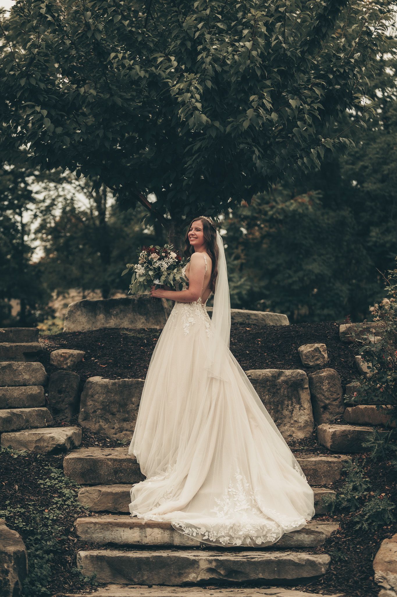 Bride in lace wedding gown poses elegantly on stone steps in garden setting.