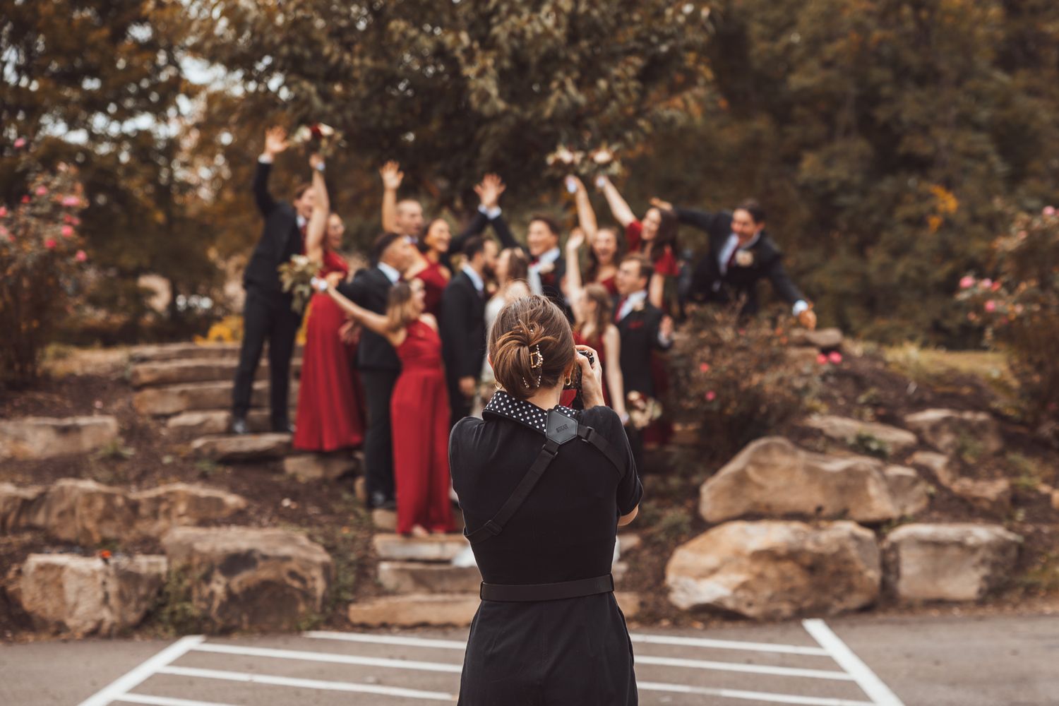A couple in formal attire dances together outdoors surrounded by autumn foliage and onlookers in red dresses.