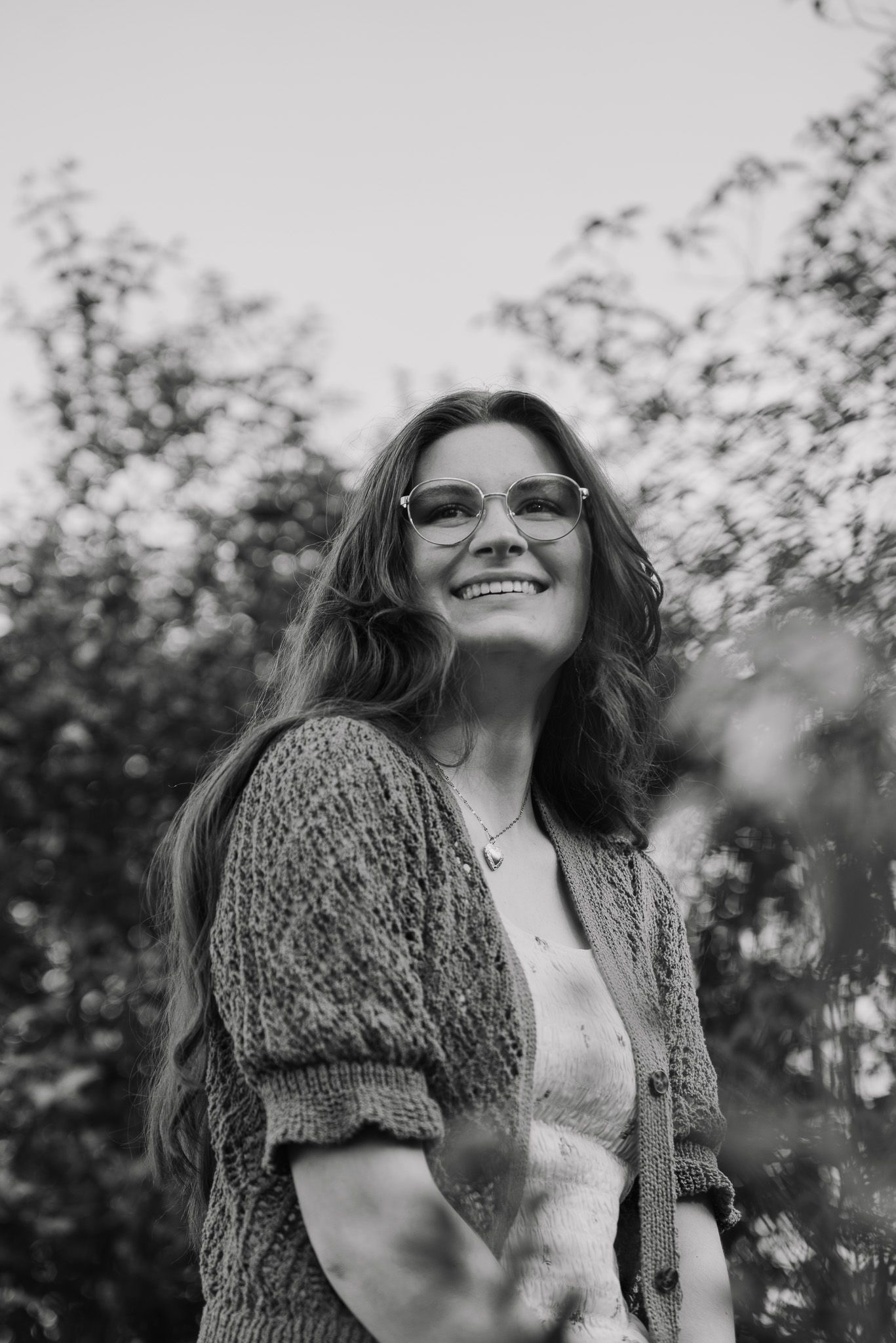 Portrait of someone wearing glasses and a knit cardigan smiling outdoors in black and white.