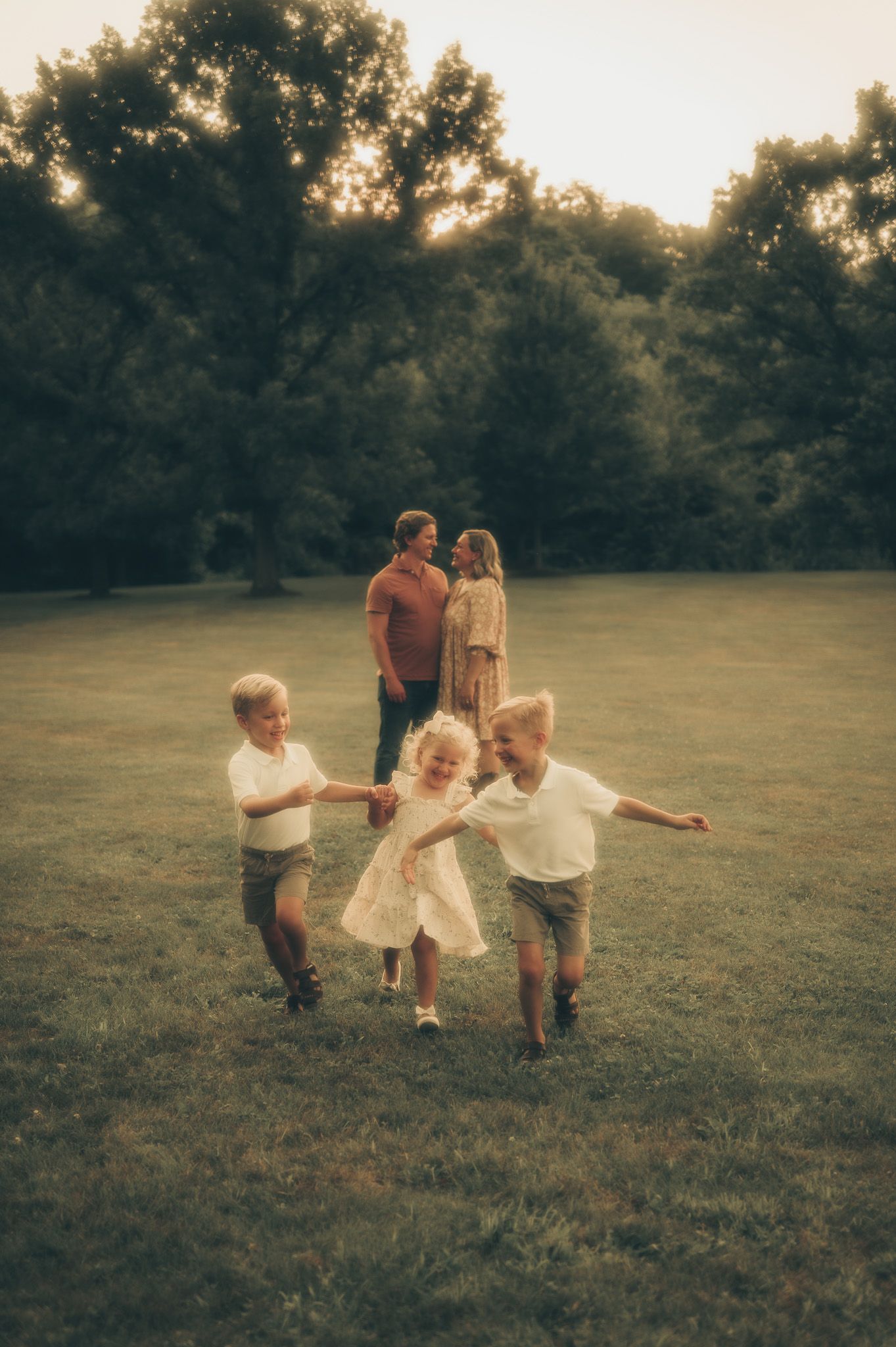 Family photo at sunset with two parents standing together while three young children play and hold hands on a green lawn.