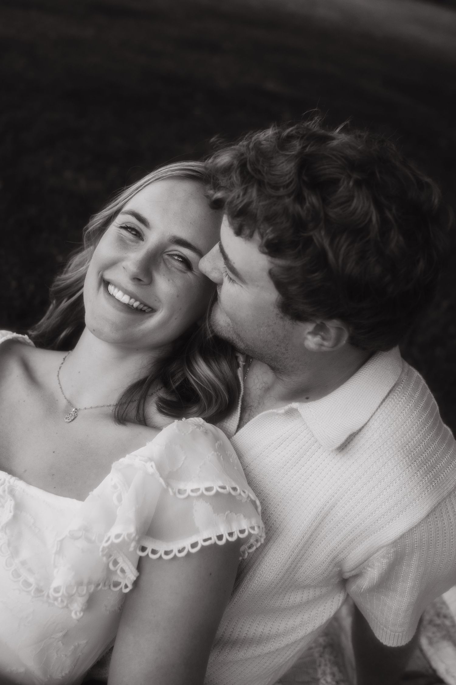 Black and white photo of a smiling woman as a curly-haired man kisses her cheek outdoors.