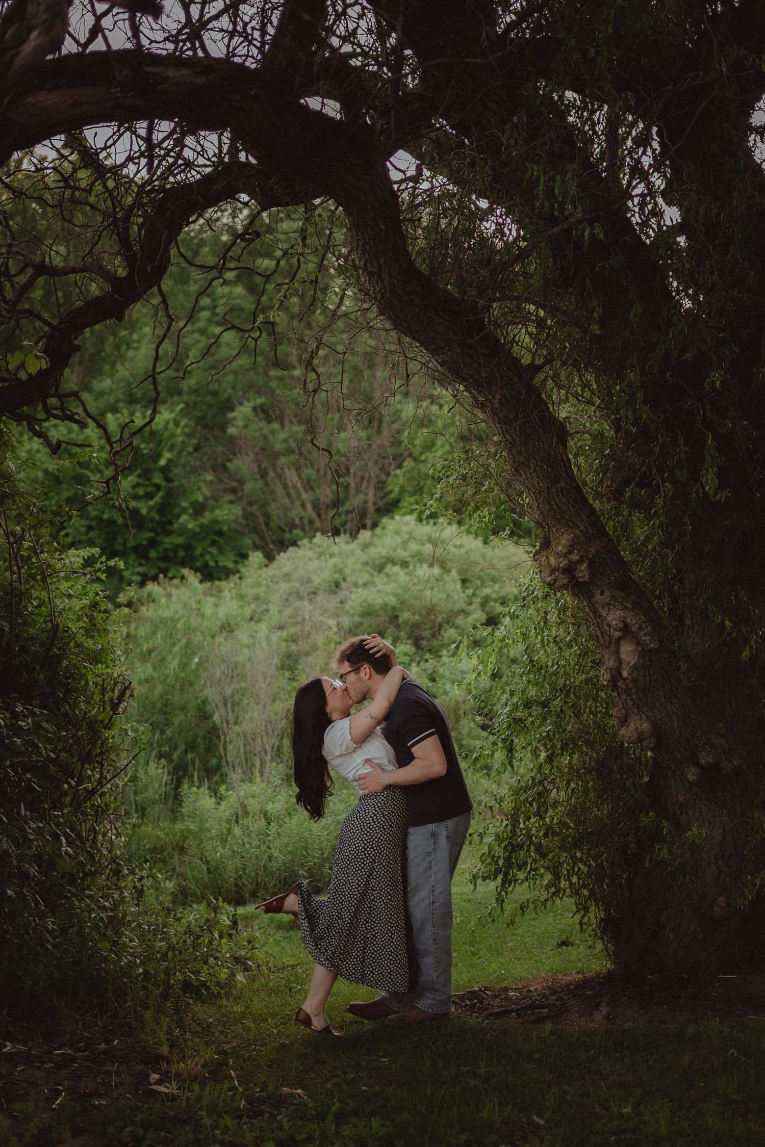 A couple shares a romantic kiss under lush green trees in a serene outdoor garden setting.